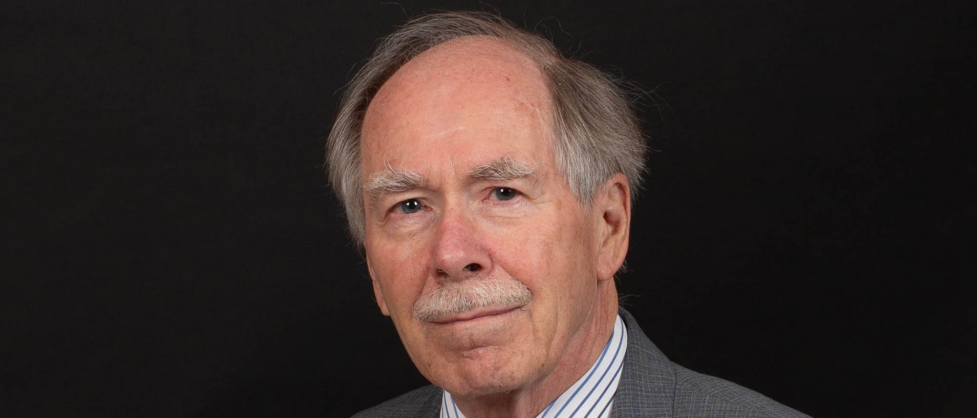 Gerard 't Hooft, ein älterer Mann mit Schnurrbart in einem grauen Anzug, blickt vor einem dunklen Hintergrund in die Kamera.