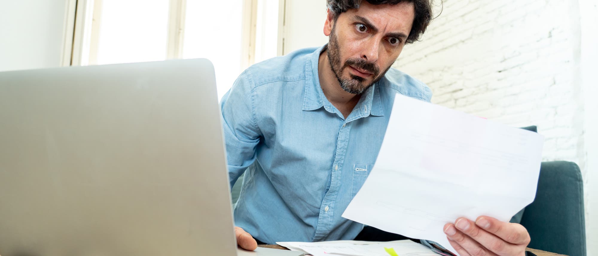 Ein Mann in einem hellblauen Jeanshemd sitzt an einem Holztisch und schaut mit weit aufgerissenen Augen auf ein Dokument in seiner Hand. Vor ihm steht ein Laptop, und auf dem Tisch liegen weitere Papiere. Im Hintergrund sind ein Fenster und eine weiße Backsteinwand zu sehen.