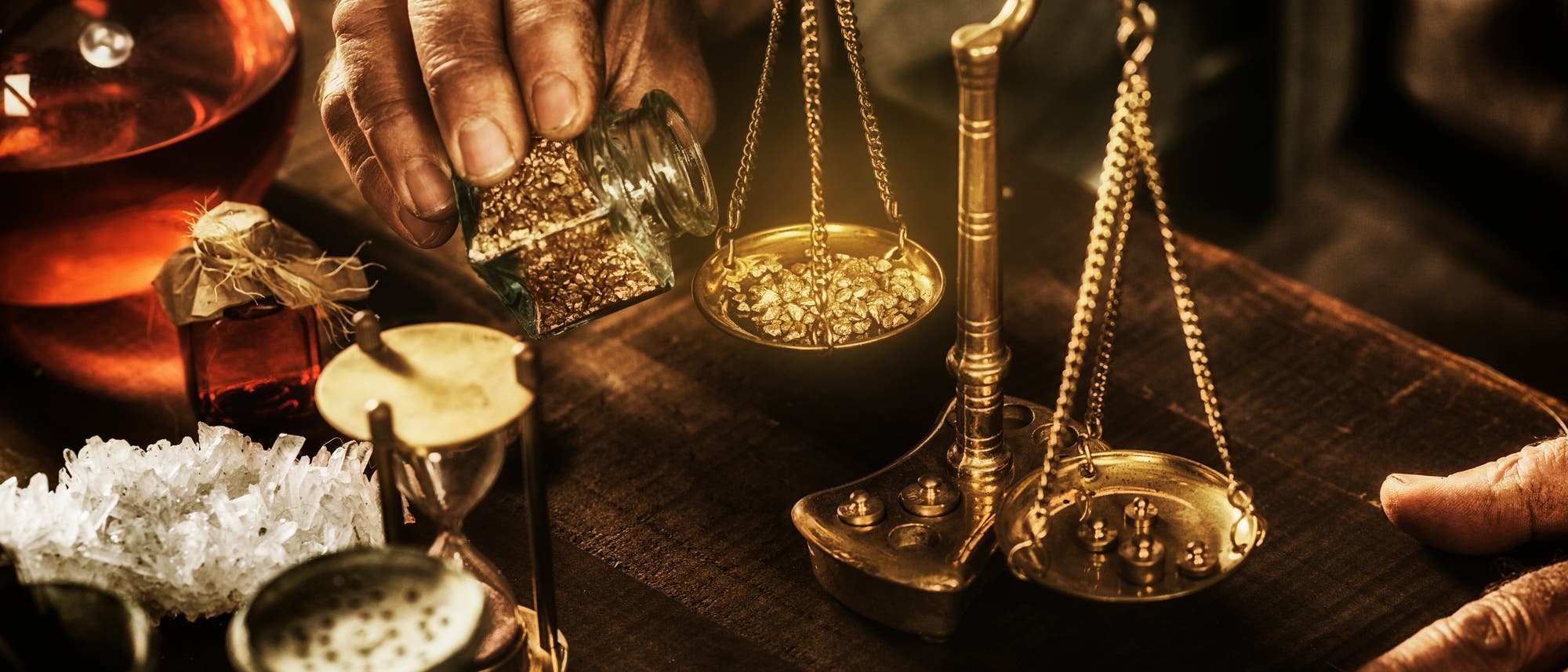 Eine Hand hält ein Glas mit goldenen Nuggets und gießt sie auf eine antike Waage auf einem Holztisch. Neben der Waage stehen eine Sanduhr, ein Glas mit roter Flüssigkeit und Kristalle. Die Szene erinnert an alchemistische Praktiken. Eine Hand hält ein Glas mit goldenen Nuggets und gießt sie auf eine antike Waage auf einem Holztisch. Neben der Waage stehen eine Sanduhr, ein Glas mit roter Flüssigkeit und Kristalle. Die Szene erinnert an alchemistische Praktiken.