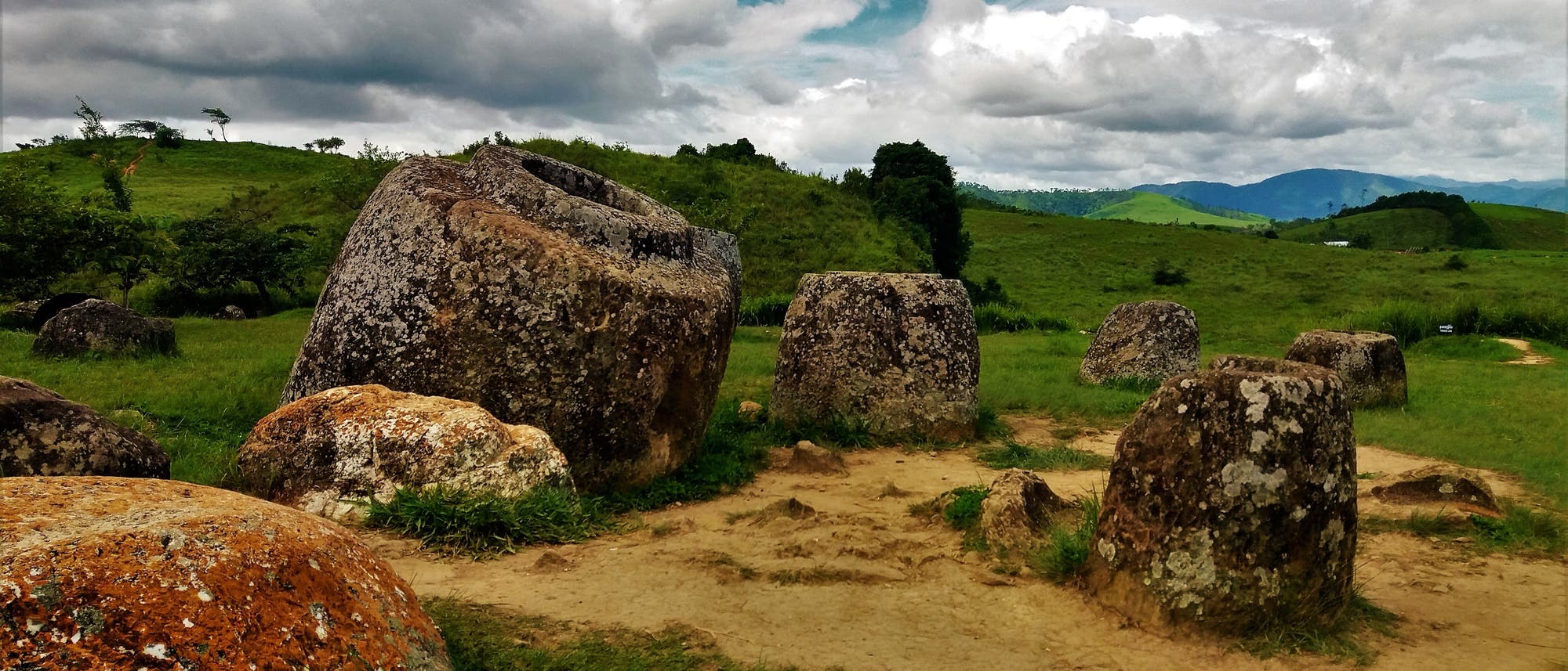 Eine Landschaft mit gro&szlig;en, antiken Steinkr&uuml;gen, die auf einer gr&uuml;nen Wiese verteilt sind. Der Himmel ist bew&ouml;lkt, und im Hintergrund sind H&uuml;gel und B&auml;ume zu sehen. Die Steinkr&uuml;ge sind unterschiedlich gro&szlig; und haben eine raue, verwitterte Oberfl&auml;che.