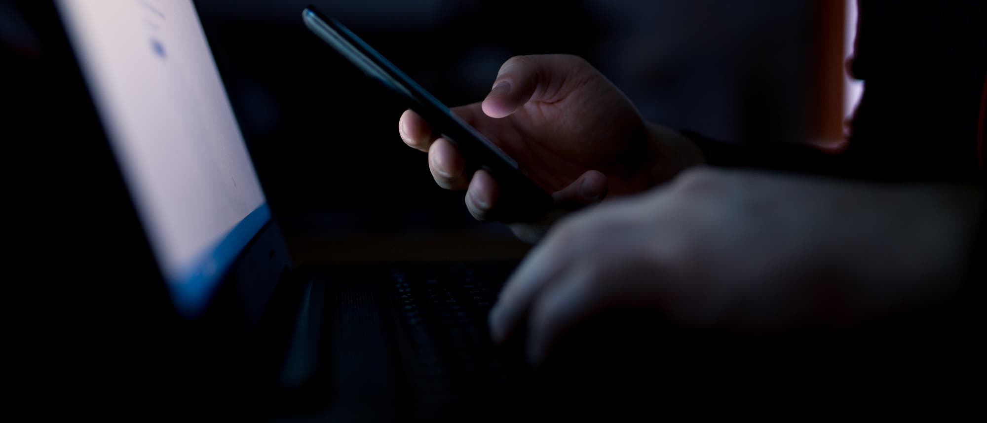 Eine Person sitzt in einem dunklen Raum vor einem Laptop und hält ein Smartphone in der Hand. Der Bildschirm des Laptops leuchtet schwach und beleuchtet die Hände der Person. Die Szene vermittelt eine Atmosphäre von Konzentration und Technologie.