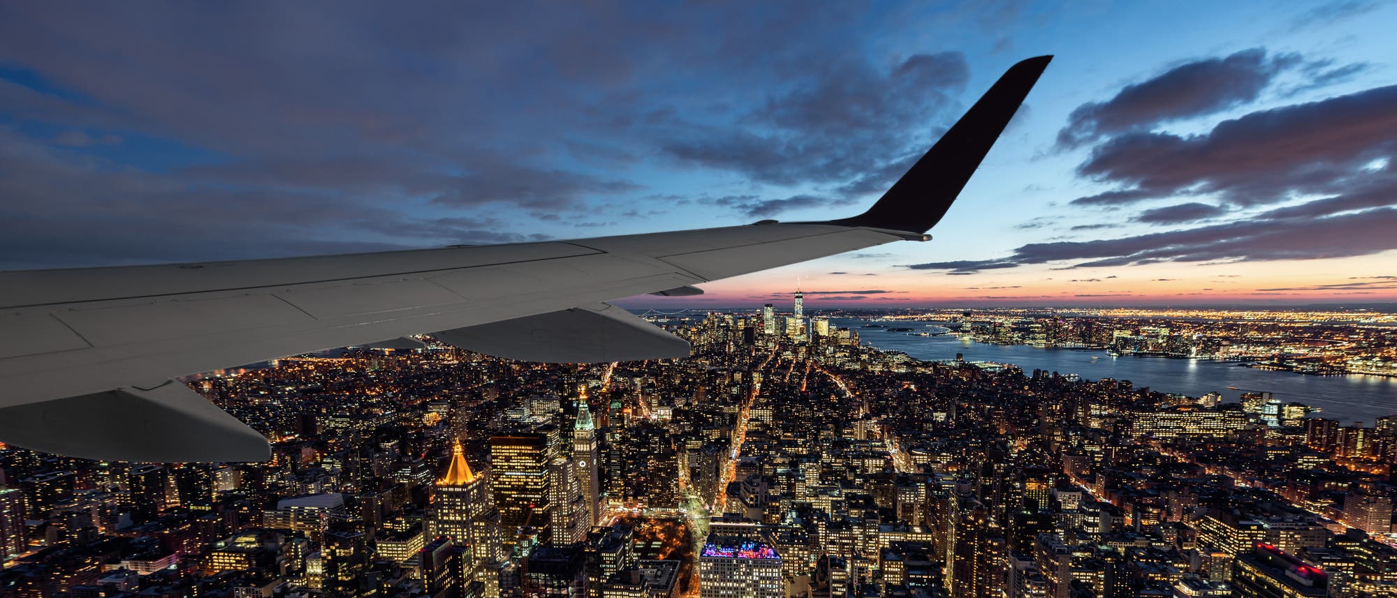 Blick aus einem Flugzeugfenster auf eine beleuchtete Stadtlandschaft bei Sonnenuntergang. Die Flügelspitze des Flugzeugs ist im Vordergrund sichtbar, während sich darunter ein dichtes Netz aus erleuchteten Straßen und Gebäuden erstreckt. Der Himmel zeigt eine Mischung aus Blau- und Orangetönen, und ein Fluss ist am Horizont zu erkennen.