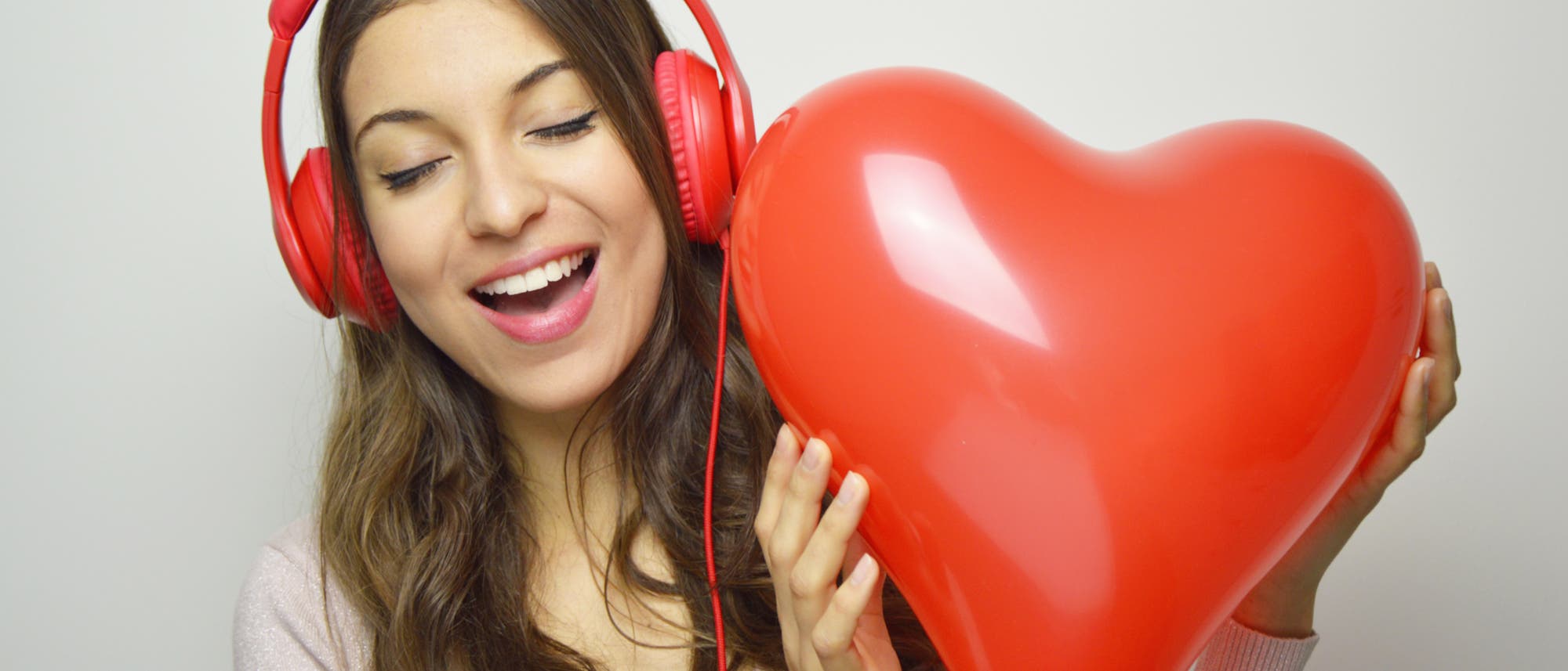 Eine Person trägt rote Kopfhörer und hält einen herzförmigen roten Ballon. Sie lächelt mit geschlossenen Augen, was Freude und Entspannung ausdrückt. Der Hintergrund ist neutral, was den Fokus auf die Person und den Ballon lenkt.