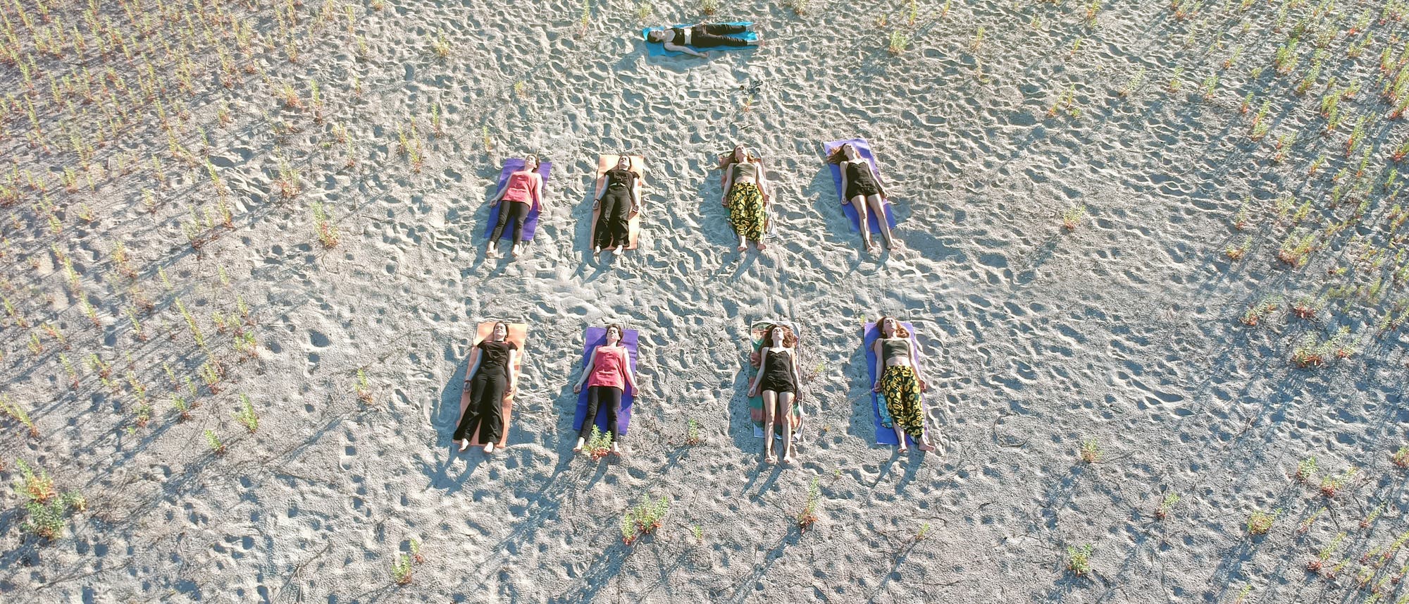 Eine Gruppe von Menschen liegt auf dem Rücken auf einem sandigen Boden, verteilt in zwei Reihen. Sie sind auf bunten Matten positioniert, umgeben von spärlichem Pflanzenwuchs. Die Szene wirkt ruhig und entspannt, möglicherweise Teil einer Yoga- oder Meditationsübung im Freien. Die Schatten der Personen sind lang und deuten auf eine tief stehende Sonne hin.