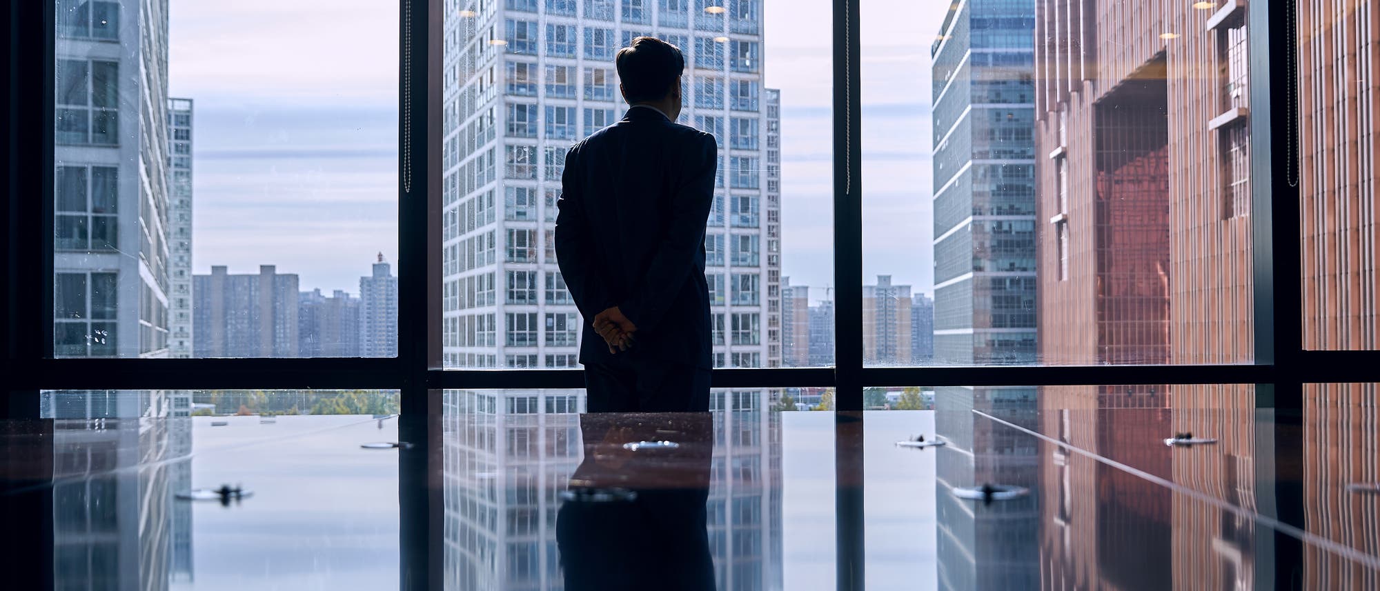 Eine Person in formeller Kleidung steht mit dem Rücken zur Kamera und blickt aus einem großen Fenster auf eine städtische Skyline mit modernen Hochhäusern. Die Reflexion der Person und der Gebäude ist auf einem glänzenden Tisch sichtbar. Die Szene vermittelt einen Eindruck von Nachdenklichkeit und urbaner Geschäftigkeit. Eine Person in formeller Kleidung steht mit dem Rücken zur Kamera und blickt aus einem großen Fenster auf eine städtische Skyline mit modernen Hochhäusern. Die Reflexion der Person und der Gebäude ist auf einem glänzenden Tisch sichtbar. Die Szene vermittelt einen Eindruck von Nachdenklichkeit und urbaner Geschäftigkeit.