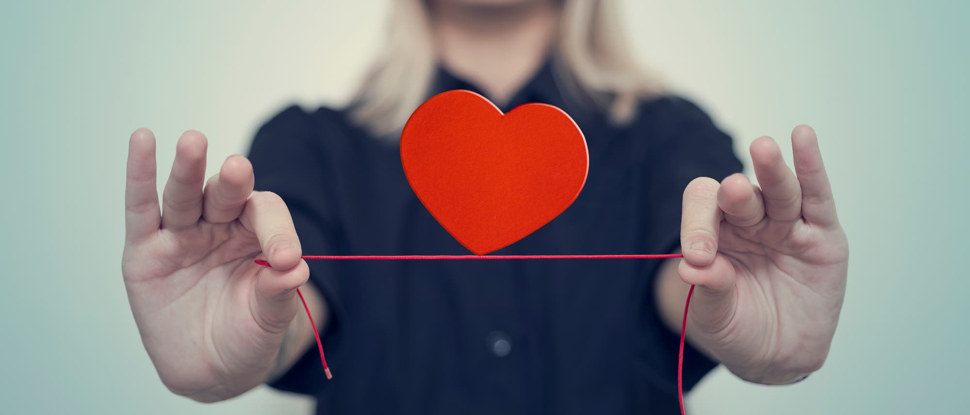 Eine Frau h&auml;lt einen roten Faden an beiden Seiten fest, auf dem ein Papierherz balanciert. Der Hintergrund ist unscharf, und der Fokus liegt auf dem Herz.