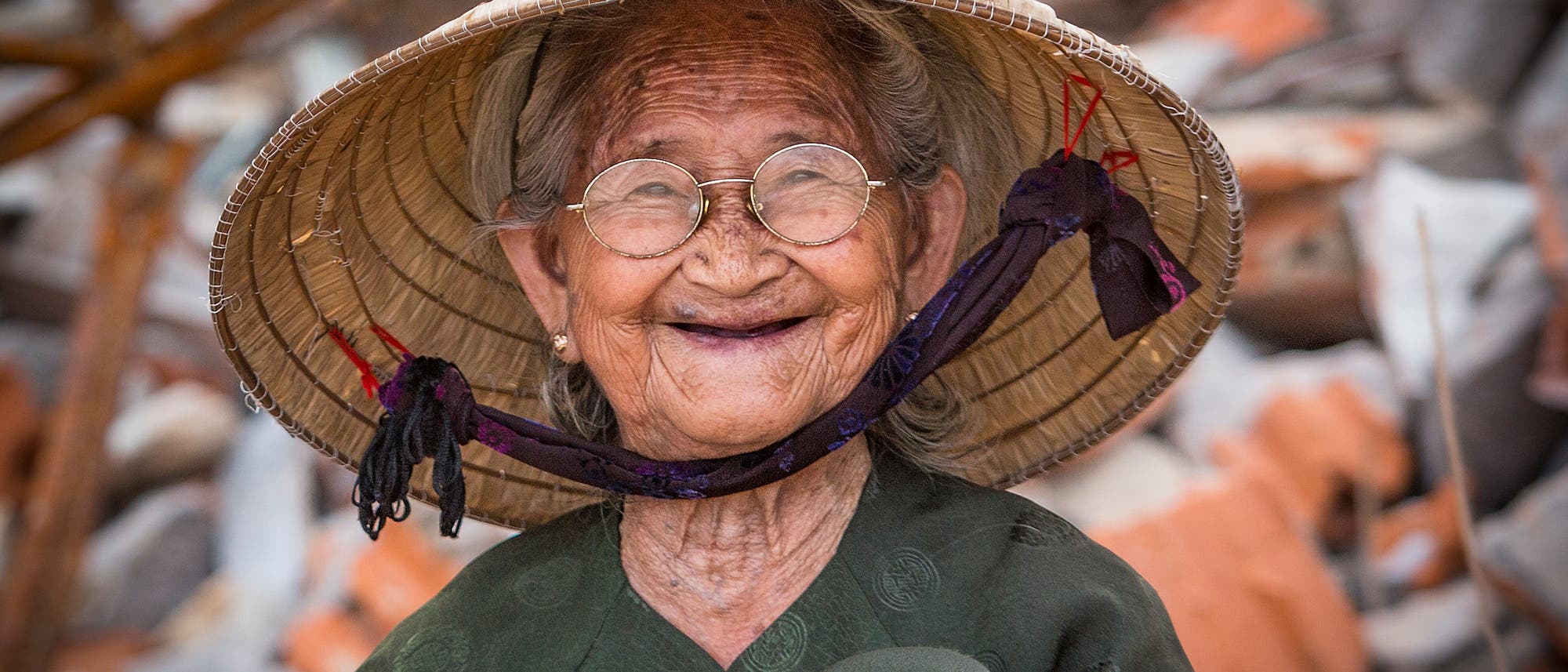 Eine lächelnde Person mit einem traditionellen, konischen Strohhut sitzt vor einem Hintergrund aus unscharfen Trümmern. Die Person trägt eine Brille und ein dunkles, gemustertes Oberteil. Der Ausdruck der Person wirkt freundlich und zufrieden. Eine lächelnde Person mit einem traditionellen, konischen Strohhut sitzt vor einem Hintergrund aus unscharfen Trümmern. Die Person trägt eine Brille und ein dunkles, gemustertes Oberteil. Der Ausdruck der Person wirkt freundlich und zufrieden.