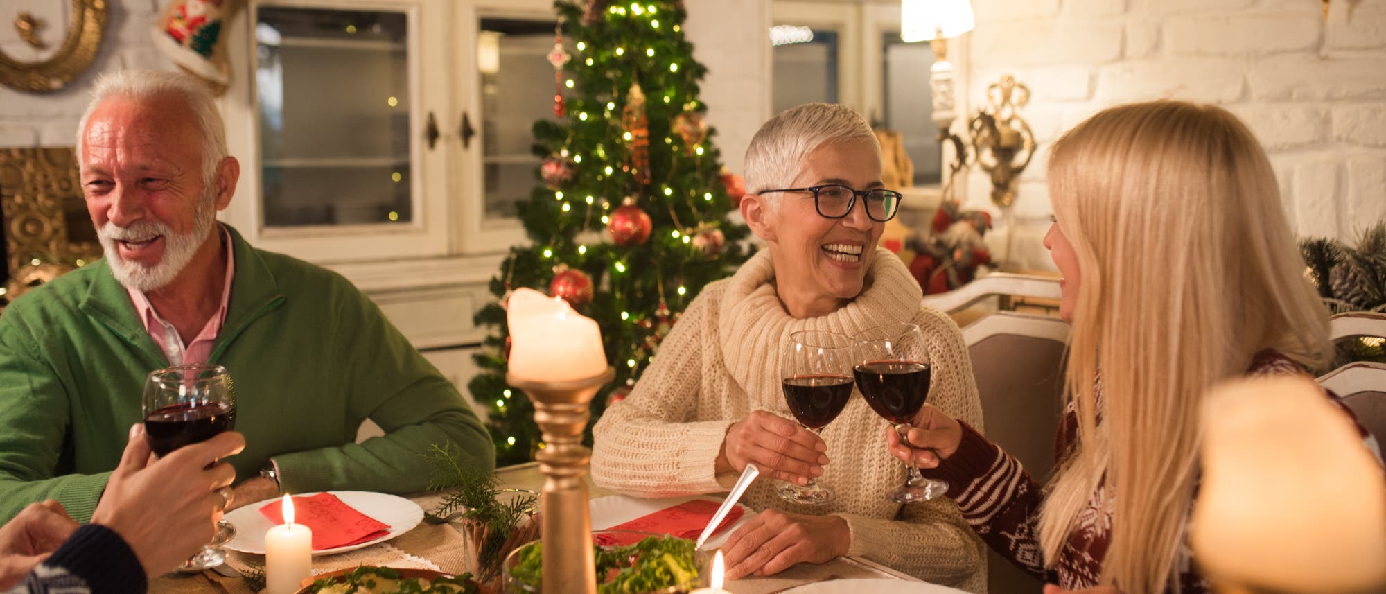 Eine Gruppe von Menschen sitzt an einem festlich gedeckten Tisch und st&ouml;&szlig;t mit Rotweingl&auml;sern an. Im Hintergrund steht ein geschm&uuml;ckter Weihnachtsbaum. Der Tisch ist mit Kerzen, Salat und verschiedenen Speisen dekoriert. Die Szene vermittelt eine warme und fr&ouml;hliche Atmosph&auml;re.