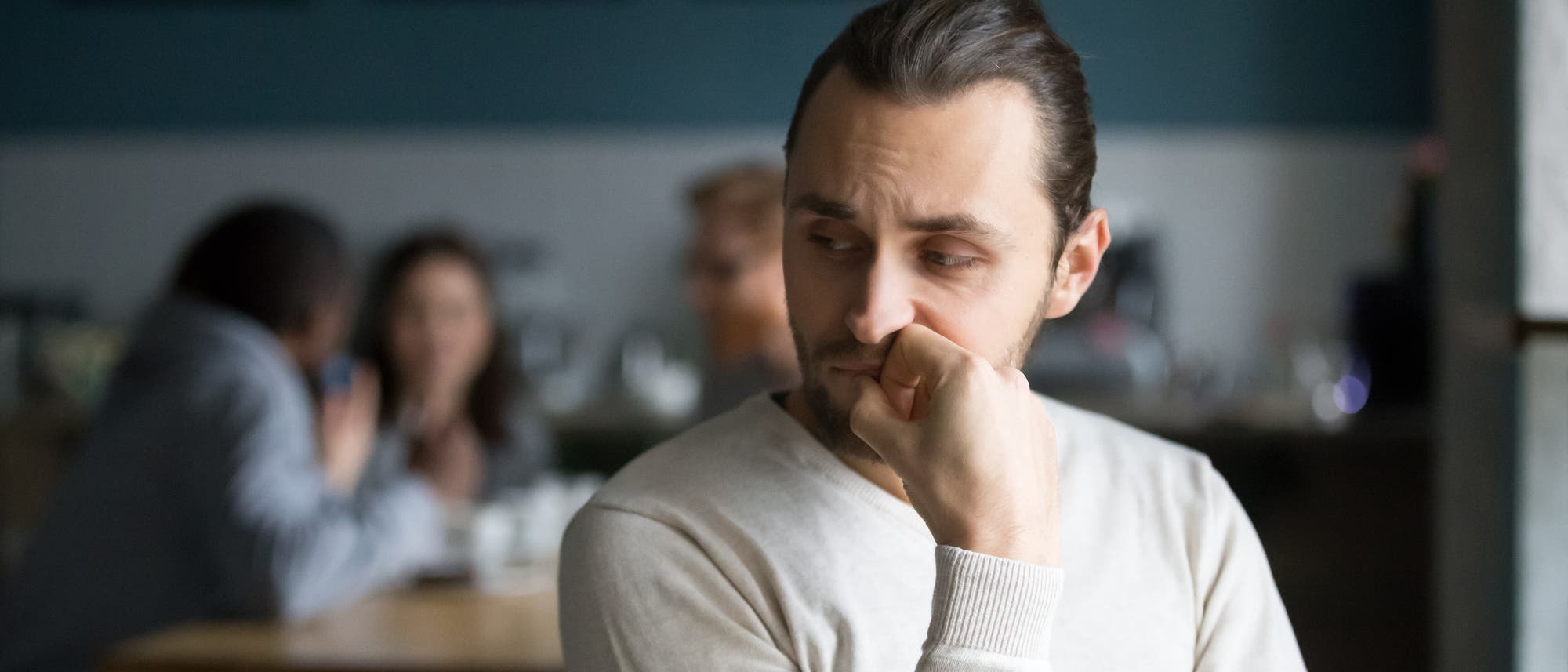 Ein etwas unglücklich dreinschauender Mann sitzt im Vordergrund. Er hat seinen Kopf auf eine Hand gestützt und leicht zur Seite gedreht, als würde er den Personen im Hintergrund lauschen, die dort im Gespräch an einem Tisch sitzen.