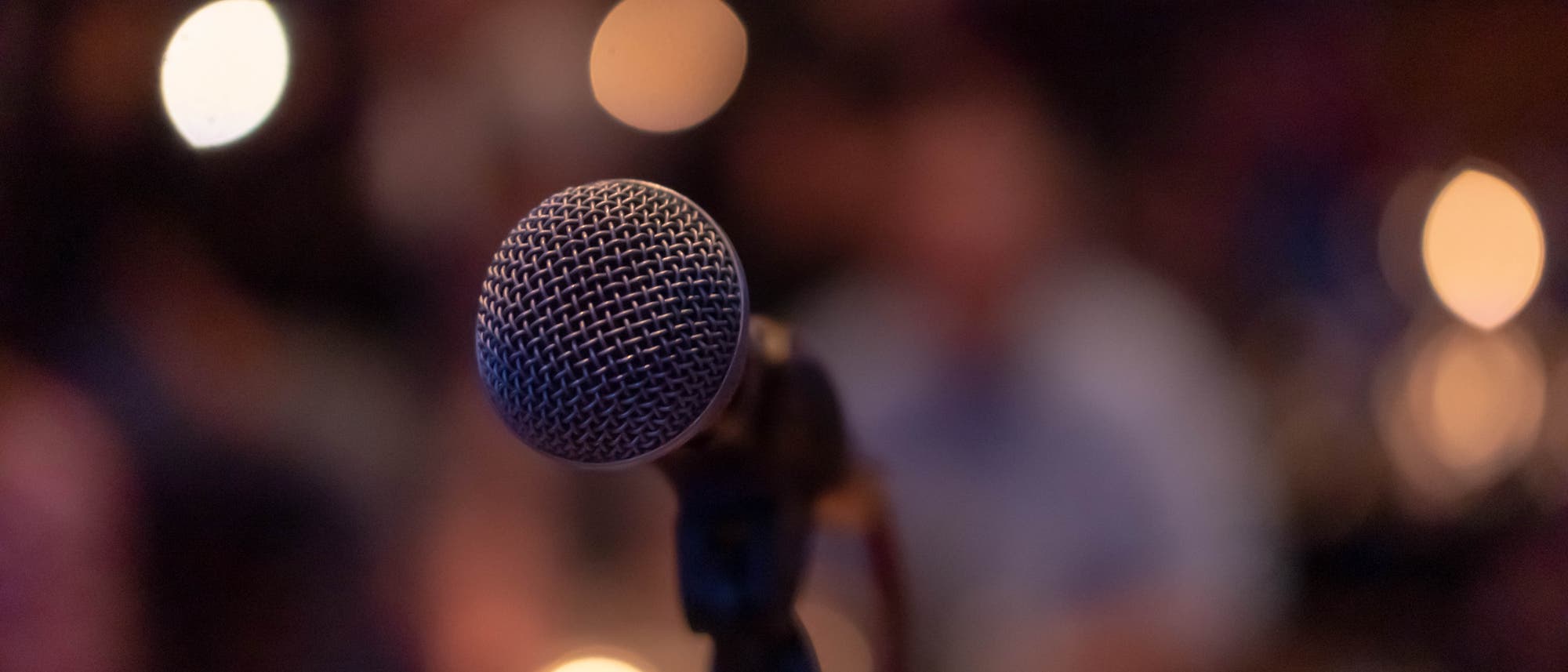 Ein Mikrofon im Vordergrund, scharf fokussiert, mit einem unscharfen Hintergrund, der warme Lichter und schemenhafte Umrisse von Menschen zeigt. Die Szene vermittelt eine Atmosphäre von Live-Performance oder öffentlichem Sprechen. Ein Mikrofon im Vordergrund, scharf fokussiert, mit einem unscharfen Hintergrund, der warme Lichter und schemenhafte Umrisse von Menschen zeigt. Die Szene vermittelt eine Atmosphäre von Live-Performance oder öffentlichem Sprechen.