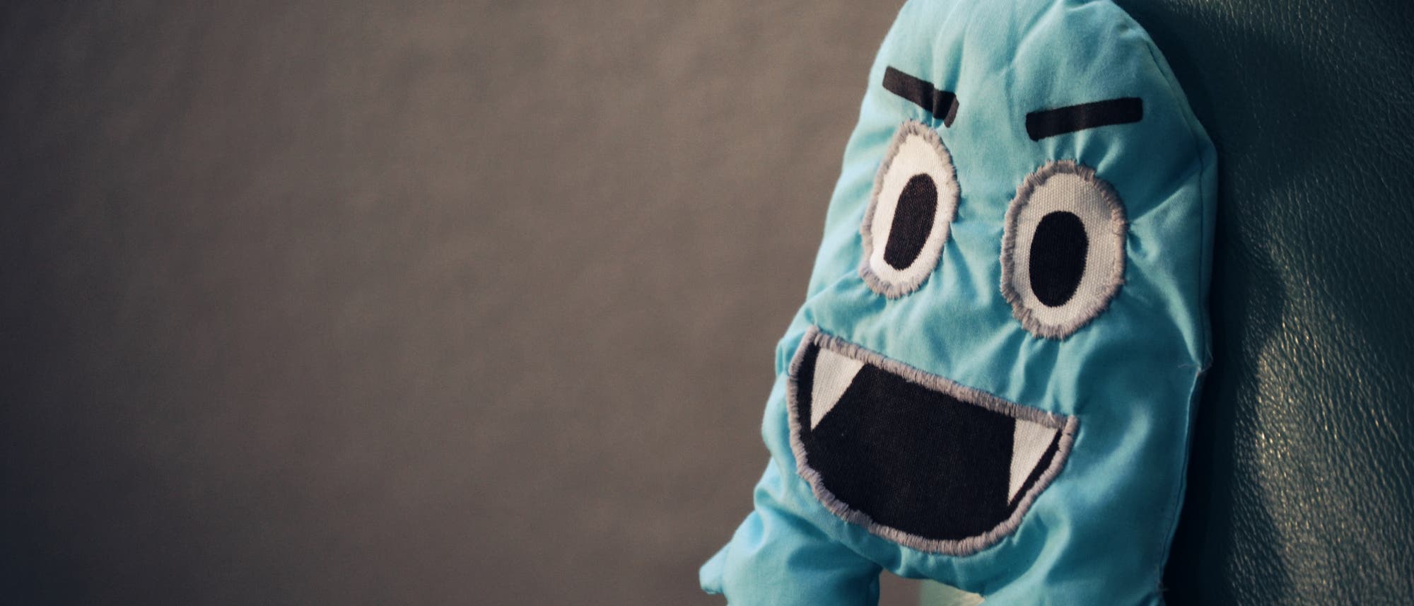 Ein blaues Stoffspielzeug mit gro&szlig;en, aufgen&auml;hten Augen und einem breiten Mund sitzt auf einem gr&uuml;nen Ledersofa. Das Spielzeug hat eine cartoonartige Erscheinung mit aufgestickten Augenbrauen und Z&auml;hnen. Der Hintergrund ist unscharf und dunkel, was den Fokus auf das Spielzeug lenkt.