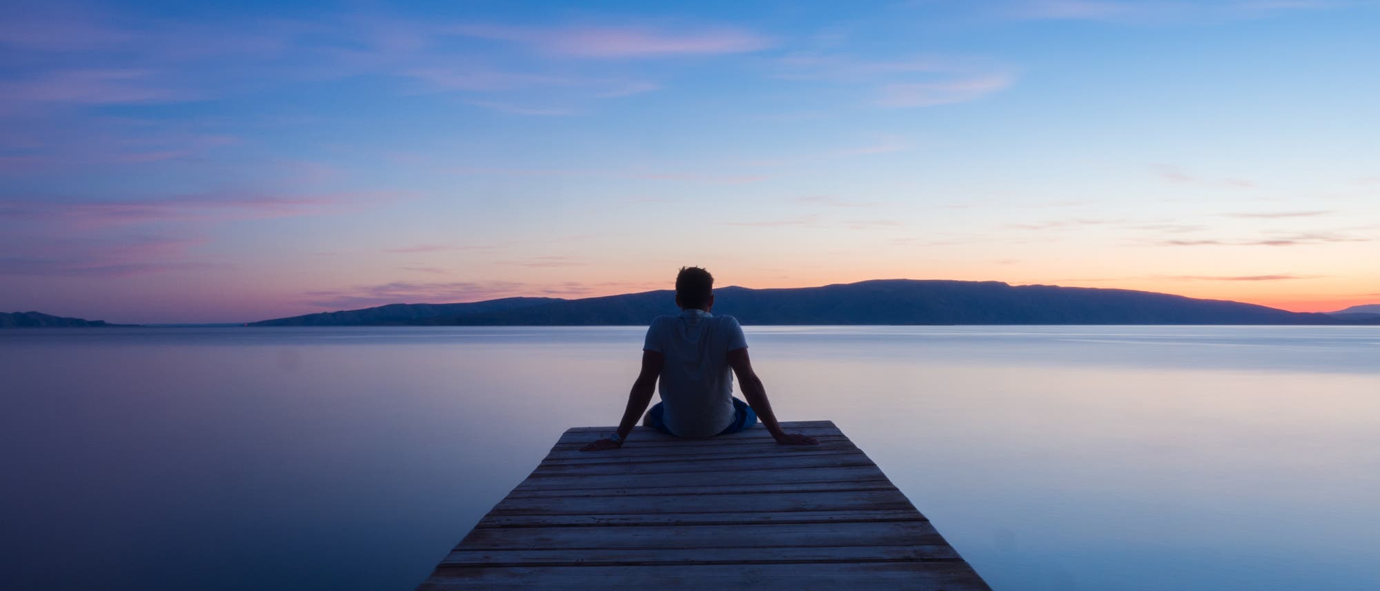 Eine Person sitzt auf einem Holzsteg, der in einen ruhigen See führt. Der Himmel zeigt ein sanftes Farbenspiel aus Blau- und Rosatönen während des Sonnenuntergangs. Im Hintergrund sind silhouettierte Hügel zu sehen. Die Szene vermittelt Ruhe und Gelassenheit. Eine Person sitzt auf einem Holzsteg, der in einen ruhigen See führt. Der Himmel zeigt ein sanftes Farbenspiel aus Blau- und Rosatönen während des Sonnenuntergangs. Im Hintergrund sind silhouettierte Hügel zu sehen. Die Szene vermittelt Ruhe und Gelassenheit.