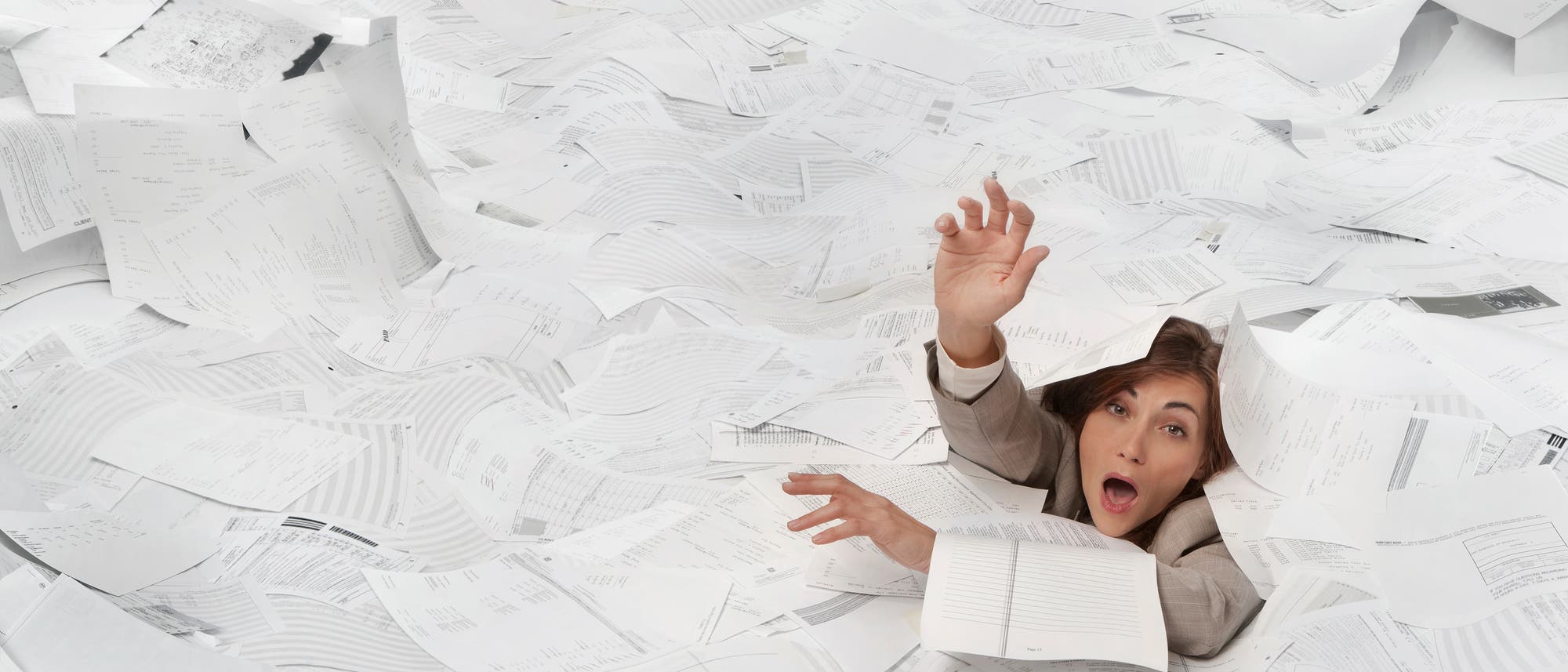 Eine Person in einem Anzug ist von einem gro&szlig;en Stapel Papier umgeben und streckt verzweifelt die Hand nach oben, als ob sie Hilfe sucht. Die Szene vermittelt ein Gef&uuml;hl von &Uuml;berforderung durch B&uuml;roarbeit oder Papierkram. Die Papiere sind unorganisiert und bedecken den gesamten Hintergrund.