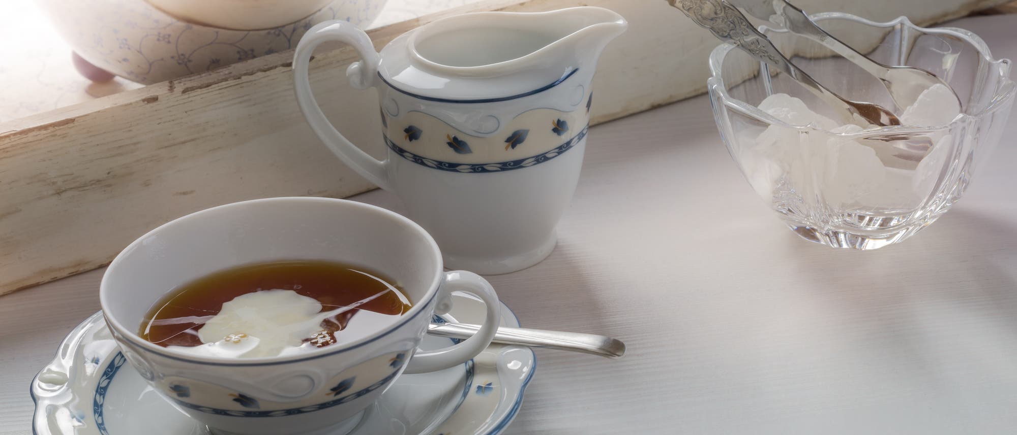 Eine elegante Teetasse mit Untertasse, gef&uuml;llt mit Tee und Sahnew&ouml;lkchen, steht auf einem Tisch. Daneben befindet sich ein Sahnek&auml;nnchen mit blauem Muster und eine Glasschale mit Kandis und einer Zange. Im Hintergrund ist ein Teil eines Teeservices zu sehen.