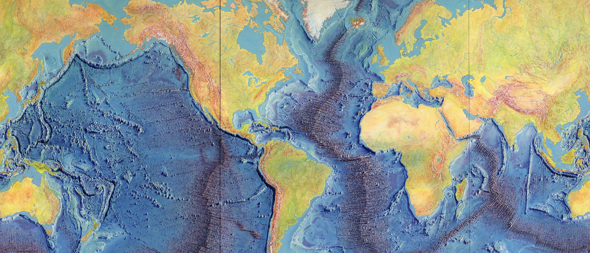 Eine physische Weltkarte zeigt die Kontinente und Ozeane der Erde. Die Landmassen sind in verschiedenen Gelb- und Braunt&ouml;nen dargestellt, um H&ouml;henunterschiede anzuzeigen, w&auml;hrend die Ozeane in Blaut&ouml;nen gehalten sind, um die Tiefen zu verdeutlichen. Gebirgsketten und Unterwasserstrukturen wie Tiefseegr&auml;ben sind detailliert abgebildet. Die Karte bietet eine umfassende Darstellung der geologischen Merkmale der Erde.