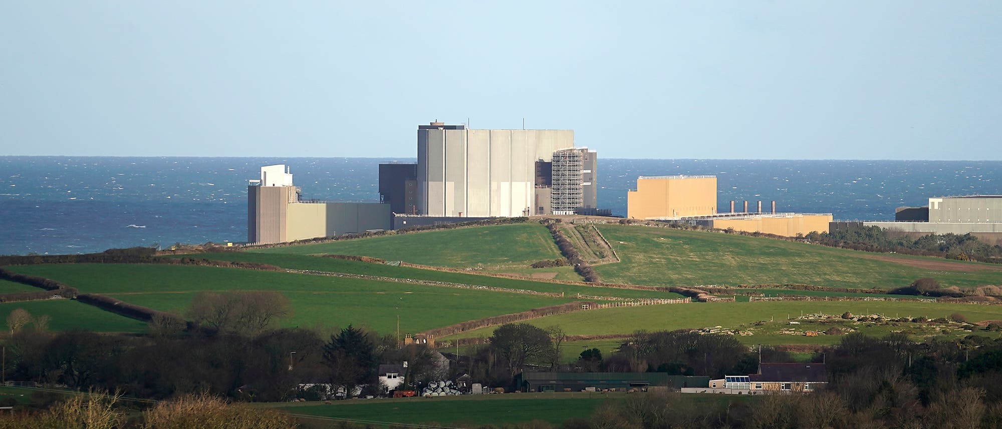 Eine Landschaft mit gr&uuml;nen Feldern und vereinzelten B&auml;umen im Vordergrund. Im Hintergrund steht ein Kernkraftwerk. Der Himmel ist klar und blau, und das Meer ist ruhig. Die Szene vermittelt eine Mischung aus nat&uuml;rlicher und industrieller Umgebung.