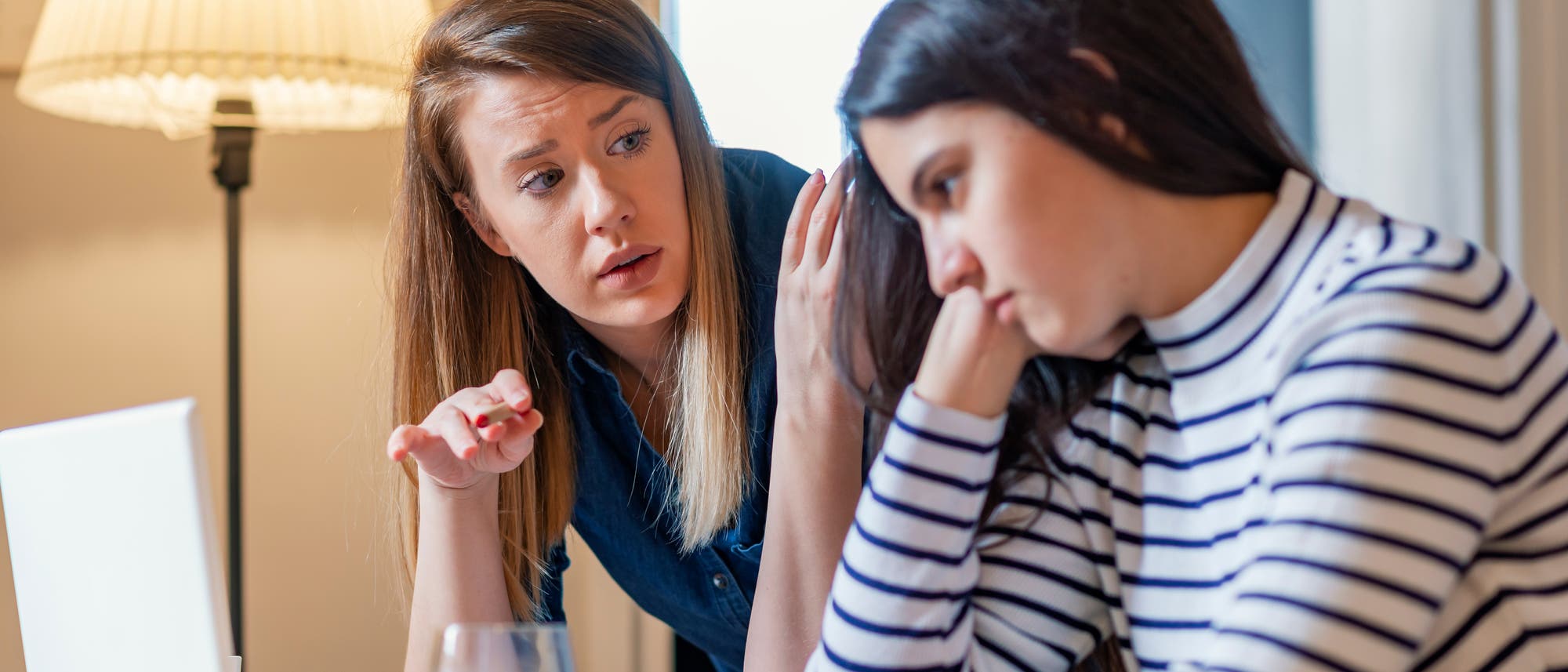 Zwei Personen sitzen an einem Tisch in einem Innenraum. Das jugendliche M&auml;dchen im Vordergrund st&uuml;tzt den Kopf auf die Hand und schaut lustlos drein. Neben ihr sitzt eine Frau in einem dunklen Hemd, beugt sich zu ihr und spricht mit ihr. Auf dem Tisch befinden sich ein Laptop, ein Glas mit Getr&auml;nk und aufgeschlagene B&uuml;cher.