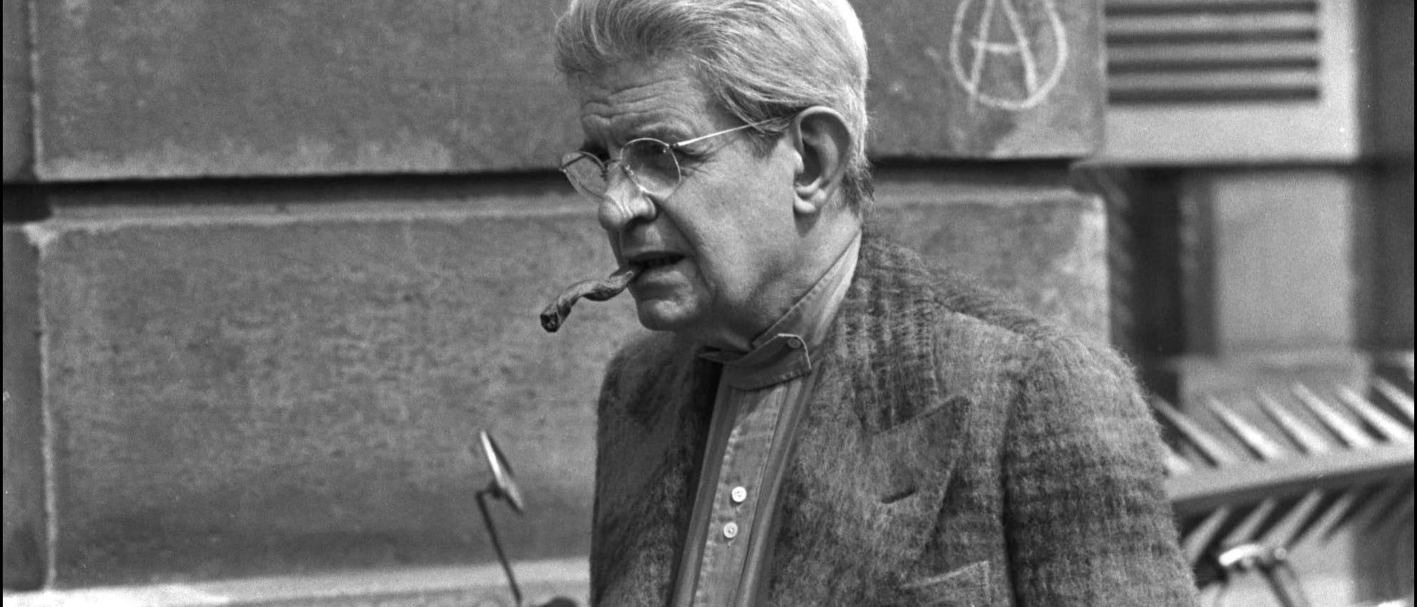 Eine Person steht vor einer Steinmauer und h&auml;lt eine Pfeife im Mund. Im Hintergrund ist ein Fahrrad zu sehen. Die Person tr&auml;gt eine Brille und einen Anzug. An der Wand ist ein Kreis mit einem "A" darin gezeichnet.