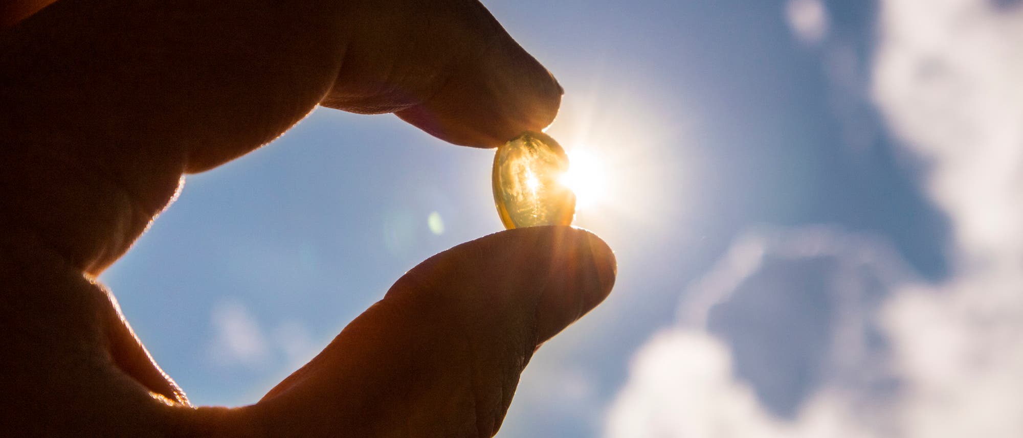 Eine Hand h&auml;lt eine durchsichtige Kapsel gegen den Himmel, durch die die Sonne scheint. Die Kapsel reflektiert das Sonnenlicht, w&auml;hrend im Hintergrund ein blauer Himmel mit einigen Wolken zu sehen ist.