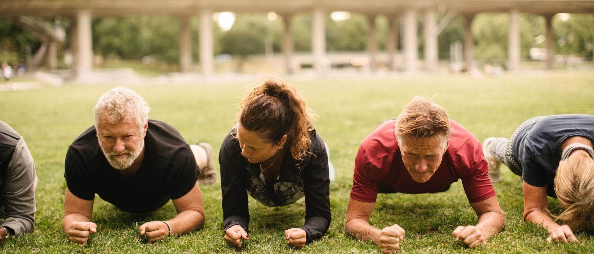 Eine Gruppe von Menschen macht Planks auf einer Wiese im Freien. Sie stützen sich auf ihre Unterarme und halten ihre Körper parallel zum Boden.Manche strengt dies mehr als andere. Eine Gruppe von Menschen macht Planks auf einer Wiese im Freien. Sie stützen sich auf ihre Unterarme und halten ihre Körper parallel zum Boden.Manche strengt dies mehr als andere.