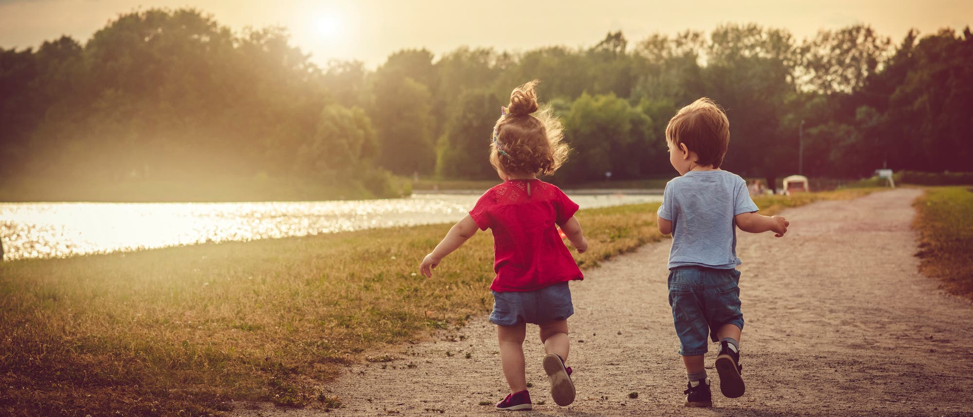 Zwei kleine Kinder laufen auf einem Schotterweg an einem Fluss in Richtung Sonnenuntergang. Das Mädchen trägt ein rotes und der Junge ein hellblaues T-Shirt. Der Himmel ist teils bewölkt und die Sonne steht tief am Horizont. Im Hintergrund sind Bäume und eine ruhige Landschaft zu sehen. Die Szene vermittelt ein Gefühl von Freiheit und Unbeschwertheit. Zwei kleine Kinder laufen auf einem Schotterweg an einem Fluss in Richtung Sonnenuntergang. Das Mädchen trägt ein rotes und der Junge ein hellblaues T-Shirt. Der Himmel ist teils bewölkt und die Sonne steht tief am Horizont. Im Hintergrund sind Bäume und eine ruhige Landschaft zu sehen. Die Szene vermittelt ein Gefühl von Freiheit und Unbeschwertheit.