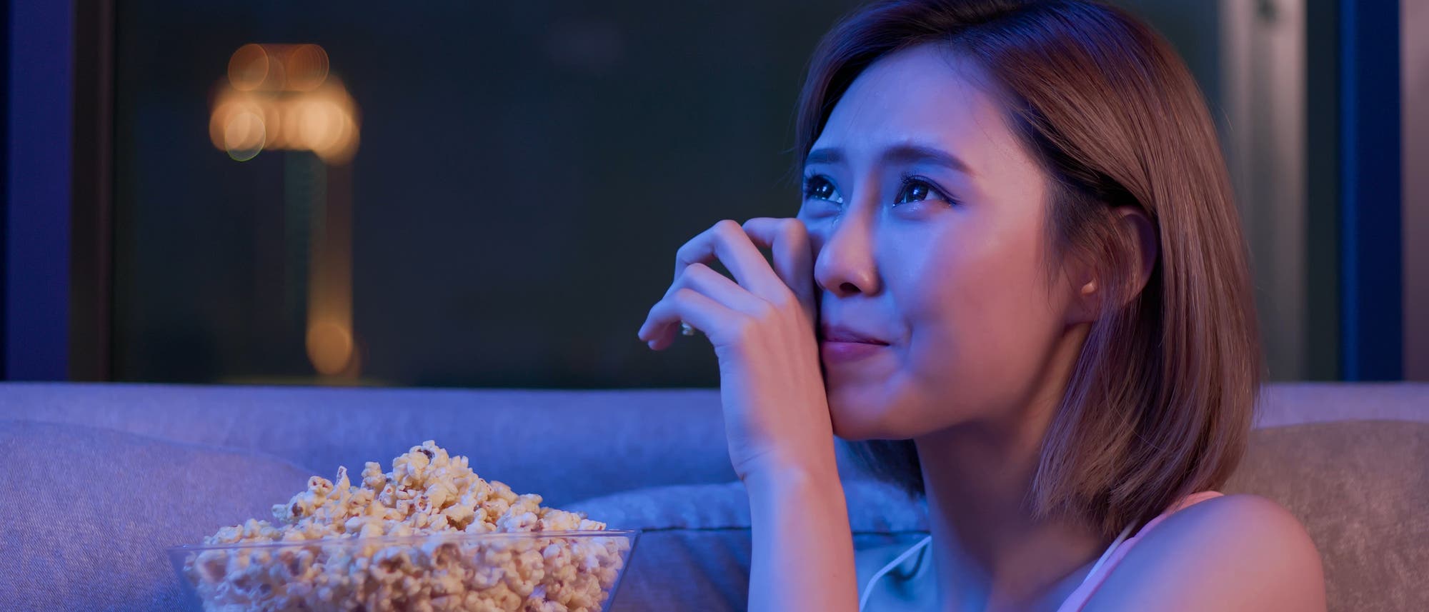 Eine Frau sitzt mit einer Sch&uuml;ssel Popcorn auf einem Sofa und schaut vermutlich einen Film. Sie wirkt emotional ber&uuml;hrt und wischt sich gerade eine Tr&auml;ne aus den Augen. Es ist dunkel im Zimmer; das Licht ist blau, der Hintergrund unscharf.