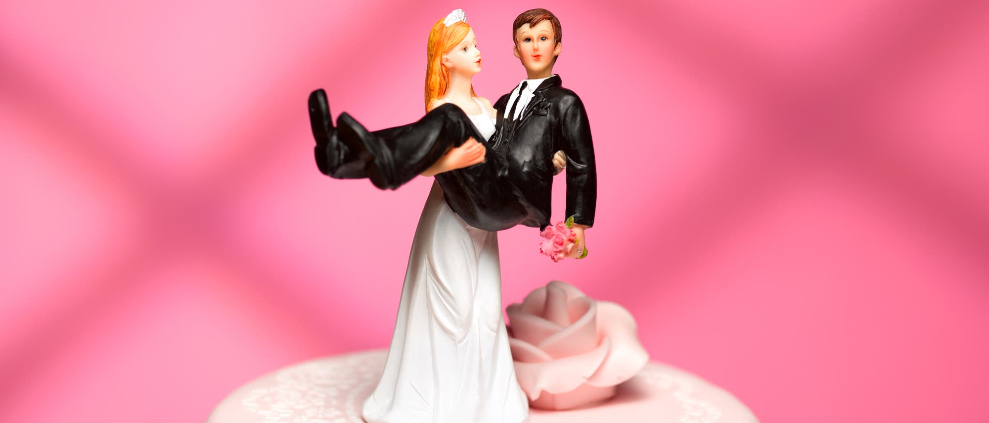 Eine Hochzeitstortenfigur zeigt eine Braut in einem weißen Kleid, die den Bräutigam in einem schwarzen Anzug trägt. Der Bräutigam hält einen kleinen Blumenstrauß. Der Hintergrund ist rosa. Die Figur steht auf einer Torte mit einer rosafarbenen Blüte. Eine Hochzeitstortenfigur zeigt eine Braut in einem weißen Kleid, die den Bräutigam in einem schwarzen Anzug trägt. Der Bräutigam hält einen kleinen Blumenstrauß. Der Hintergrund ist rosa. Die Figur steht auf einer Torte mit einer rosafarbenen Blüte.