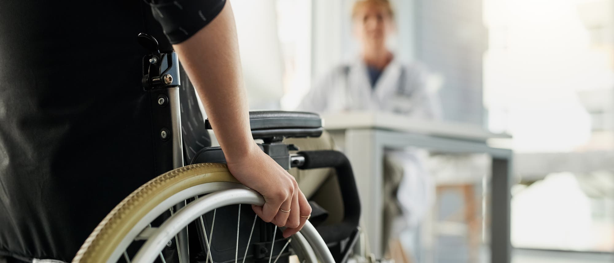 Eine Person im Rollstuhl sitzt in einem hellen Raum und hält das Rad des Rollstuhls. Im Hintergrund sitzt eine unscharf dargestellte Person in einem weißen Kittel an einem Tisch, möglicherweise ein Arzt oder medizinisches Fachpersonal. Die Szene vermittelt eine medizinische oder pflegerische Umgebung. Eine Person im Rollstuhl sitzt in einem hellen Raum und hält das Rad des Rollstuhls. Im Hintergrund sitzt eine unscharf dargestellte Person in einem weißen Kittel an einem Tisch, möglicherweise ein Arzt oder medizinisches Fachpersonal. Die Szene vermittelt eine medizinische oder pflegerische Umgebung.
