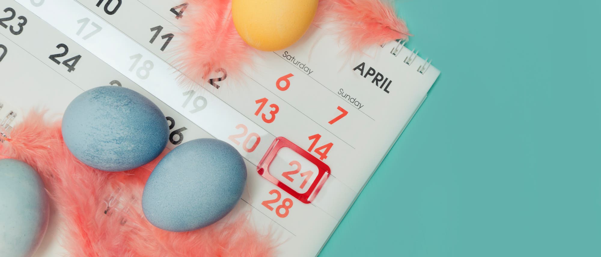 Ein Kalenderblatt f&uuml;r April 2019 liegt auf einem t&uuml;rkisfarbenen Hintergrund. Der 21. April ist rot markiert. Auf dem Kalender liegen mehrere gef&auml;rbte Ostereier, umgeben von rosa Federn. Das Bild vermittelt eine fr&uuml;hlingshafte und &ouml;sterliche Stimmung.