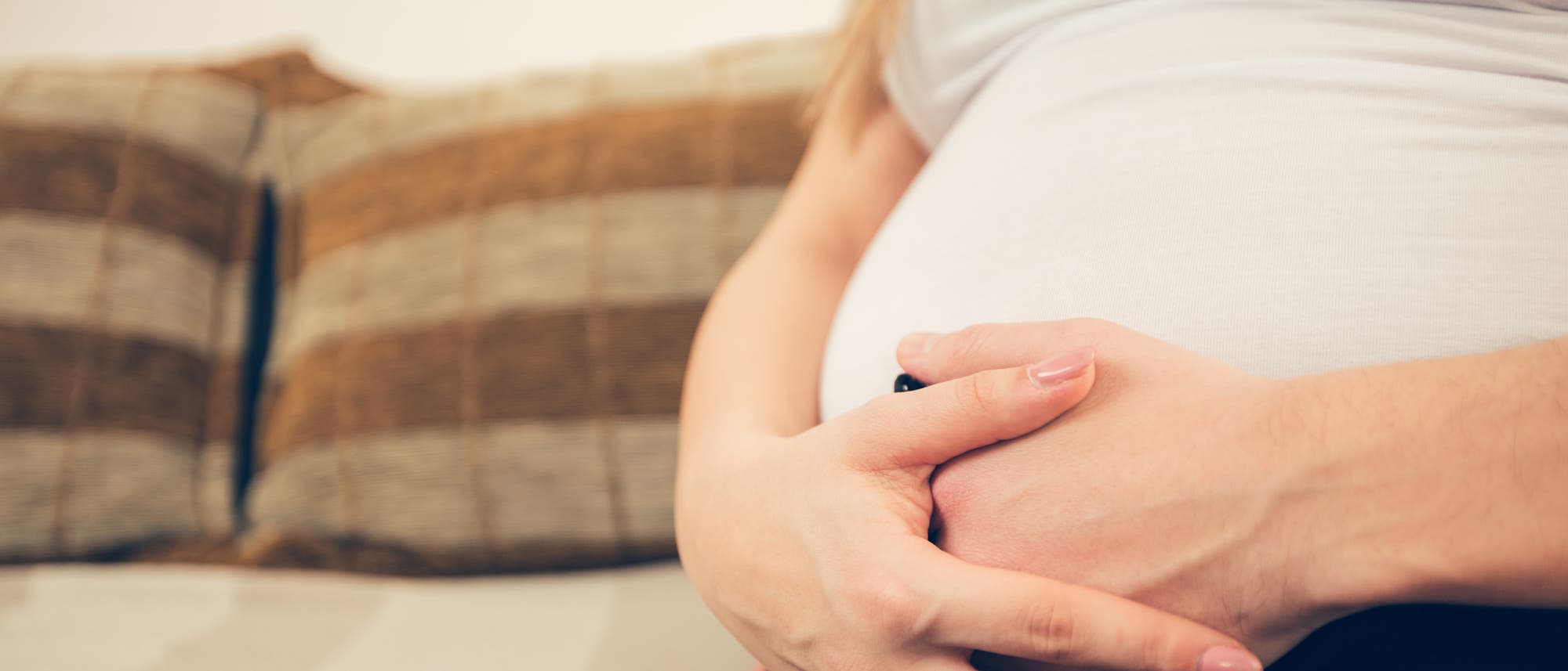 ein Foto zeigt eine Schwangere in weißem Trägertop und schwarzer Hose, die auf dem Sofa sitzt und die Hände vor dem. Babybauch verschränkt hat. Das Gesicht ist nicht zu sehen.