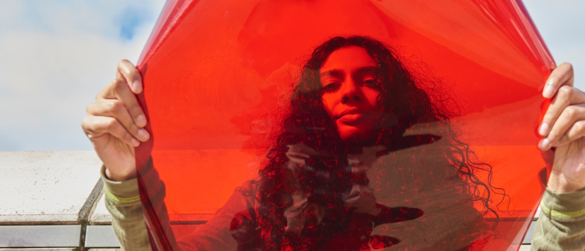Eine Person h&auml;lt eine gro&szlig;e, rote, transparente Folie vor sich, im Hintergrund ist blauer Himmel mit einigen Wolken zu sehen. Sie tr&auml;gt ein lang&auml;rmliges, helles Oberteil; die rote Folie erzeugt einen Farbeffekt auf ihrem Gesicht.