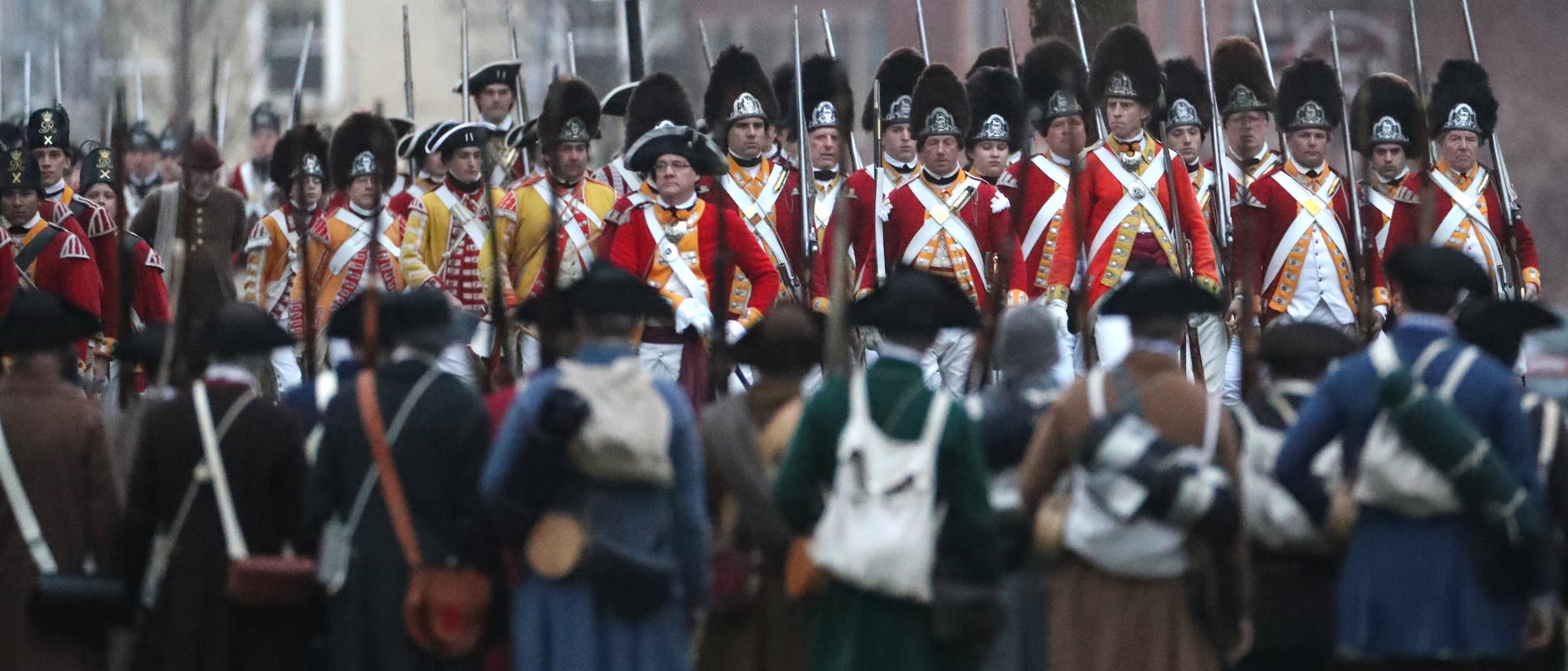 Eine Gruppe von Menschen in historischen Milit&auml;runiformen steht sich auf einem Feld gegen&uuml;ber. Im Hintergrund sind Soldaten in roten Uniformen mit wei&szlig;en Gurten und schwarzen H&uuml;ten zu sehen, die Gewehre tragen. Im Vordergrund stehen Personen in braunen und blauen Uniformen mit H&uuml;ten, die ebenfalls Gewehre tragen. Ein Schild mit der Aufschrift "Battle Green" ist im Hintergrund sichtbar. Die Szene stellt eine historische Nachstellung dar.