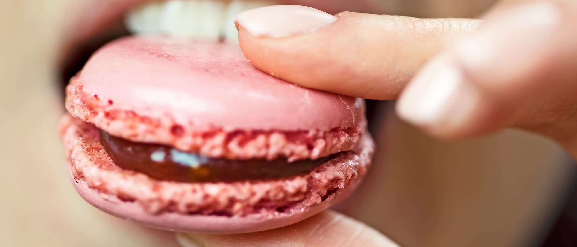 Eine Nahaufnahme eines rosa Macarons, das von einer Hand gehalten wird. Das Macaron wird zum Mund einer Person gef&uuml;hrt, wobei die F&uuml;llung zwischen den beiden H&auml;lften sichtbar ist. Die Szene vermittelt den Moment kurz vor dem Genuss des Geb&auml;cks.