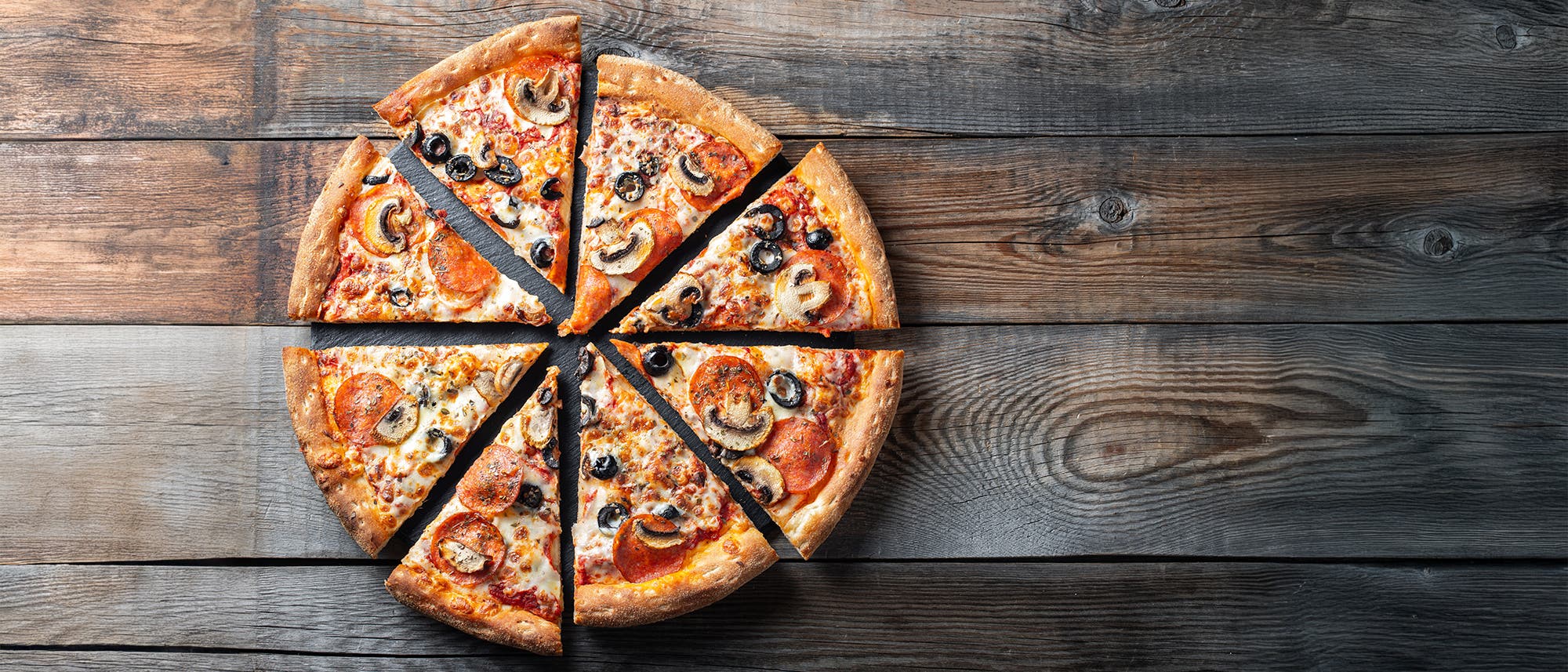 Eine Pizza in acht gleichmäßige Stücke geschnitten, mit knuspriger Kruste, belegt mit Salami, Pilzen, schwarzen Oliven und geschmolzenem Käse, liegt auf einem rustikalen Holztisch. Eine Pizza in acht gleichmäßige Stücke geschnitten, mit knuspriger Kruste, belegt mit Salami, Pilzen, schwarzen Oliven und geschmolzenem Käse, liegt auf einem rustikalen Holztisch.