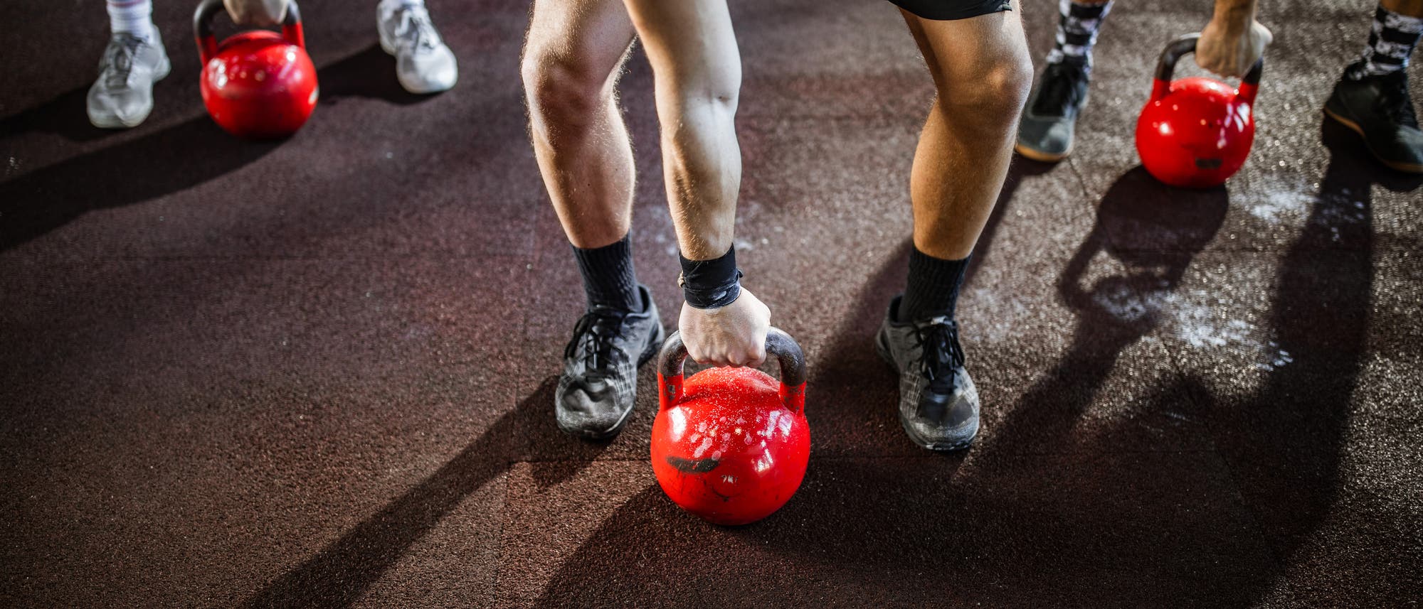 Eine Gruppe von Personen trainiert mit roten Kettlebells auf einem dunklen Boden. Die Personen sind in einer geb&uuml;ckten Haltung und heben die Kettlebells an. Sie tragen sportliche Kleidung und Turnschuhe. Die Szene vermittelt eine dynamische und konzentrierte Atmosph&auml;re.