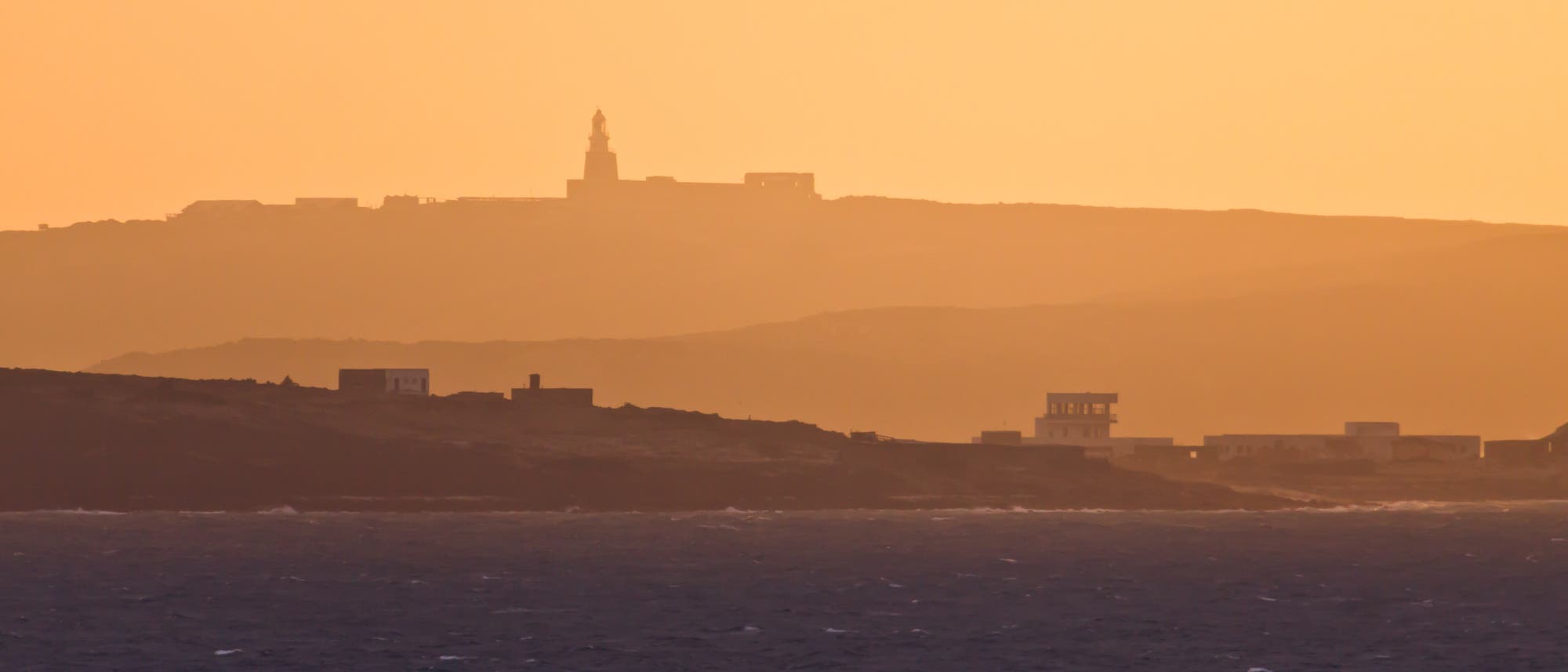 Eine Küstenlandschaft bei Sonnenuntergang. Im Vordergrund ist das Meer zu sehen, während im Hintergrund eine hügelige Landschaft mit mehreren Gebäuden, darunter ein Leuchtturm, in warmen Orangetönen beleuchtet wird. Die Szene vermittelt eine ruhige und friedliche Atmosphäre. Eine Küstenlandschaft bei Sonnenuntergang. Im Vordergrund ist das Meer zu sehen, während im Hintergrund eine hügelige Landschaft mit mehreren Gebäuden, darunter ein Leuchtturm, in warmen Orangetönen beleuchtet wird. Die Szene vermittelt eine ruhige und friedliche Atmosphäre.