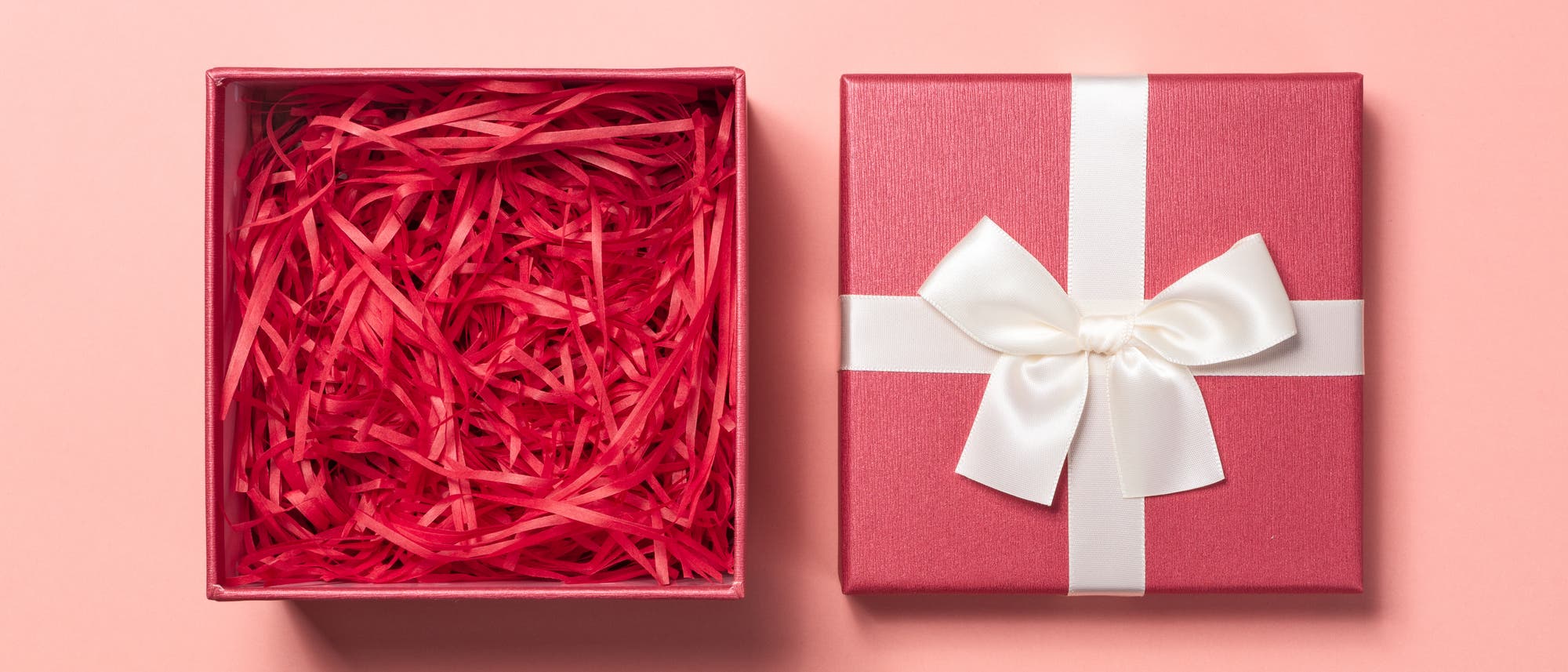Eine ge&ouml;ffnete rote Geschenkbox mit rotem F&uuml;llmaterial neben einem geschlossenen, gleichfarbigen Geschenk mit wei&szlig;er Schleife auf einem rosa Hintergrund.