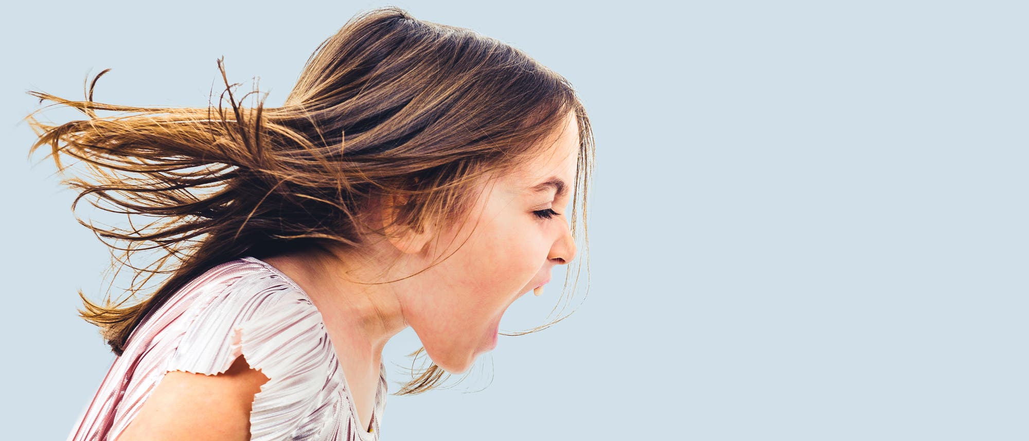 Ein Kind mit langen Haaren schreit mit weit ge&ouml;ffnetem Mund. Der Hintergrund ist einfarbig hellblau, was den Fokus auf das Gesicht und den Ausdruck des Kindes lenkt. Die Haare wehen zur Seite, was auf eine Bewegung oder einen Windsto&szlig; hindeutet.