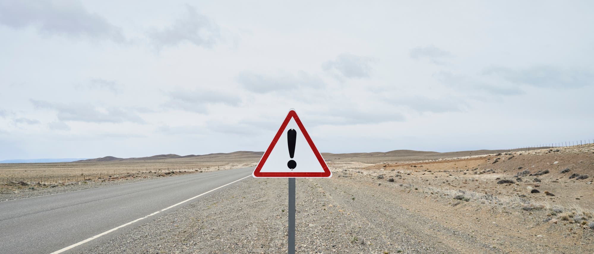 Ein dreieckiges Warnschild mit einem Ausrufezeichen steht am Rand einer einsamen, asphaltierten Stra&szlig;e in einer kargen, trockenen Landschaft. Der Himmel ist bew&ouml;lkt, und die Umgebung wirkt weitl&auml;ufig und unbewohnt. Das Schild signalisiert Vorsicht oder Gefahr auf der Stra&szlig;e.