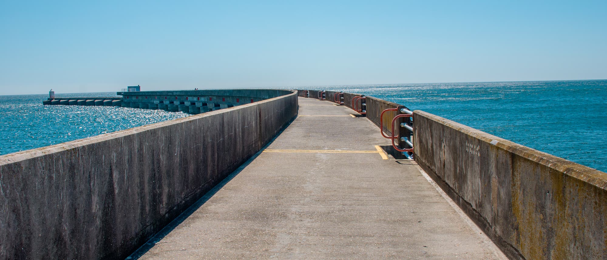Ein langer, gerader Pier aus Beton erstreckt sich ins Meer, umgeben von blauem Wasser und klarem Himmel. Auf der linken Seite ist ein kleiner Leuchtturm sichtbar. Die Sonne reflektiert auf der Wasseroberfläche, und entlang des Piers sind rote Geländer angebracht. Ein langer, gerader Pier aus Beton erstreckt sich ins Meer, umgeben von blauem Wasser und klarem Himmel. Auf der linken Seite ist ein kleiner Leuchtturm sichtbar. Die Sonne reflektiert auf der Wasseroberfläche, und entlang des Piers sind rote Geländer angebracht.