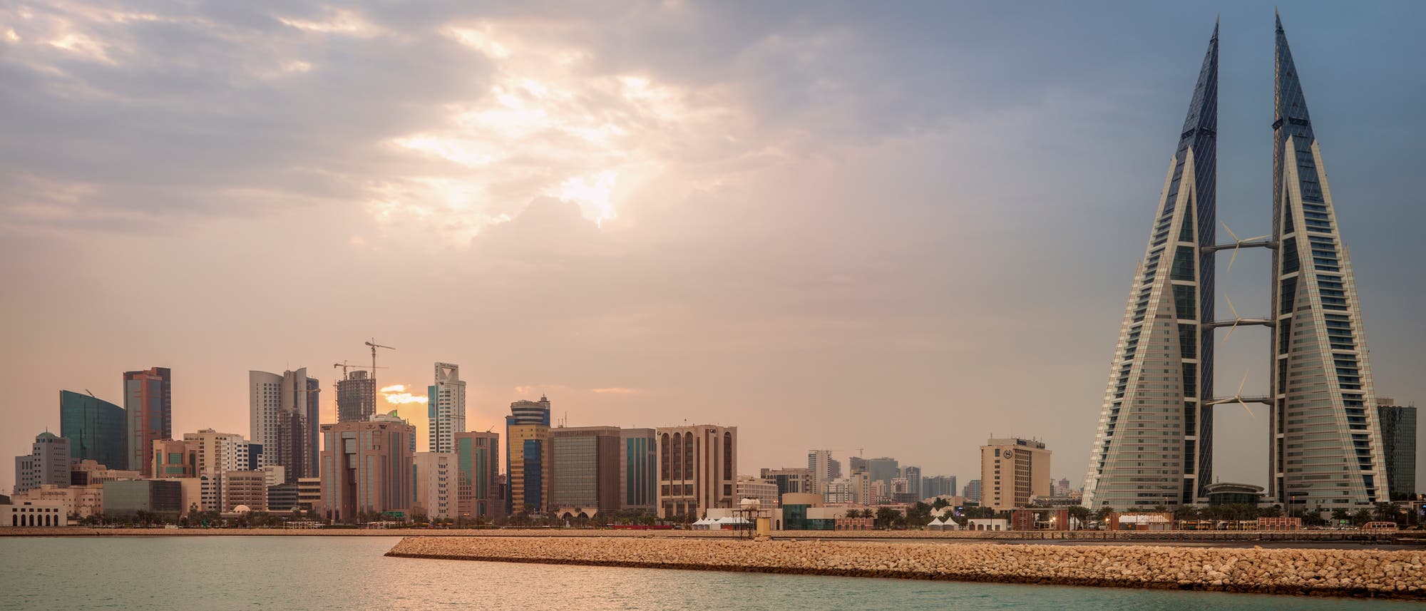 Stadtansicht von Manama, Bahrain, bei Sonnenuntergang. Im Vordergrund ist das Wasser des Persischen Golfs zu sehen, w&auml;hrend im Hintergrund moderne Hochh&auml;user die Skyline dominieren. Besonders auff&auml;llig sind die beiden spitz zulaufenden T&uuml;rme des Bahrain World Trade Centers. Der Himmel ist bew&ouml;lkt, mit Sonnenstrahlen, die durch die Wolken brechen.