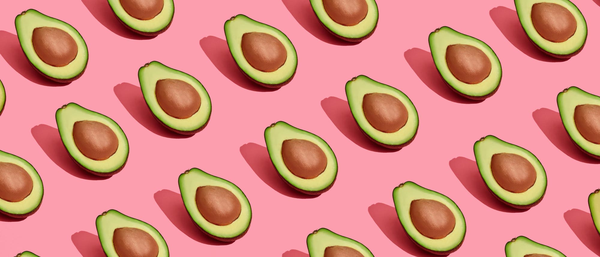 Mehrere Hälften von Avocados mit Kernen sind gleichmäßig auf einem rosa Hintergrund angeordnet. Die Avocados sind in diagonalen Reihen platziert, und jede Hälfte wirft einen Schatten. Die Komposition erzeugt ein modernes und frisches Muster. Mehrere Hälften von Avocados mit Kernen sind gleichmäßig auf einem rosa Hintergrund angeordnet. Die Avocados sind in diagonalen Reihen platziert, und jede Hälfte wirft einen Schatten. Die Komposition erzeugt ein modernes und frisches Muster.