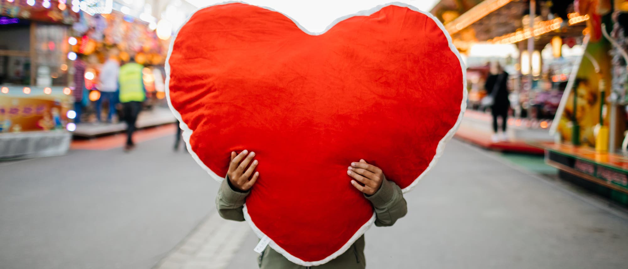 Eine Person hält ein großes, rotes, herzförmiges Kissen vor sich, so dass das Gesicht verdeckt ist. Im Hintergrund ist ein Jahrmarkt mit bunten Lichtern und Fahrgeschäften zu sehen. Die Szene vermittelt eine fröhliche und lebendige Atmosphäre.