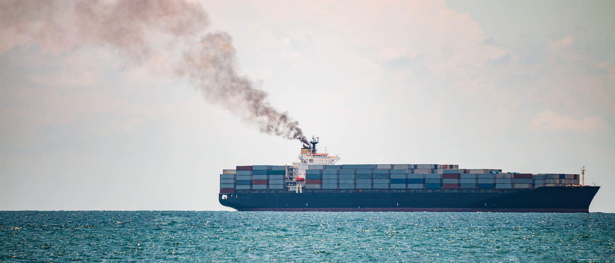 Ein Containerschiff fährt auf dem Meer, aus dem Schornstein steigt schwarzer Rauch auf.