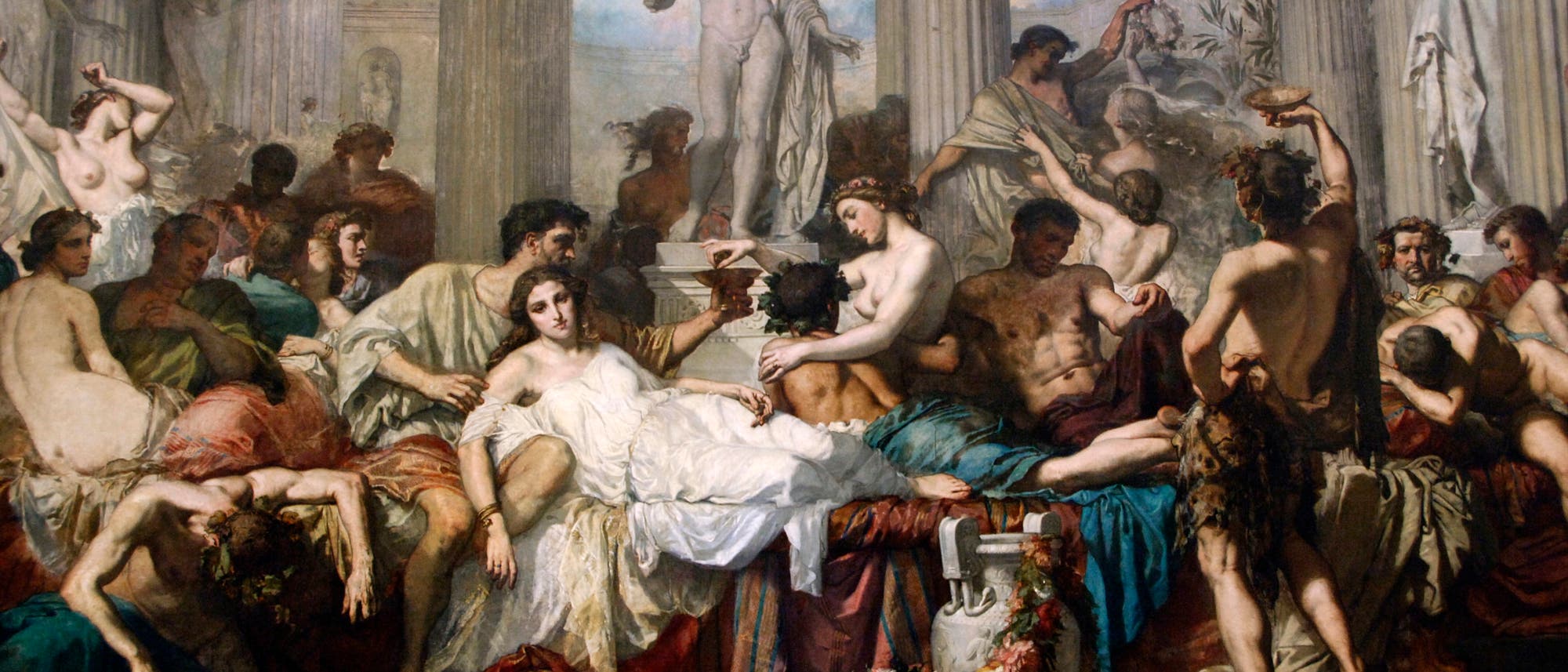 Eine klassische Gem&auml;ldeszene zeigt eine Gruppe von Menschen in einer antiken Umgebung, die an einem Bankett teilnimmt. Im Zentrum liegt eine Frau in einem wei&szlig;en Gewand auf einer Couch, umgeben von anderen Personen, die in lebhaften Gespr&auml;chen vertieft sind. Im Hintergrund steht eine gro&szlig;e Statue, die die Szene &uuml;berblickt. Die Architektur ist von S&auml;ulen und Skulpturen gepr&auml;gt, die den antiken Stil unterstreichen. Blumen und Fr&uuml;chte schm&uuml;cken den Vordergrund, was auf eine festliche Atmosph&auml;re hinweist.