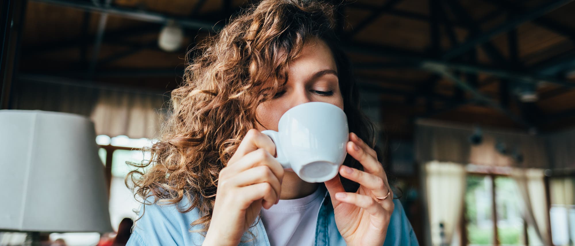 Eine Person sitzt in einem gem&uuml;tlichen Caf&eacute; und genie&szlig;t einen Schluck aus einer wei&szlig;en Tasse. Sie tr&auml;gt ein hellblaues Hemd und eine wei&szlig;e Uhr. Im Hintergrund sind Holzbalken und gro&szlig;e Fenster zu sehen, die nat&uuml;rliches Licht hereinlassen. Die Atmosph&auml;re wirkt entspannt und einladend.