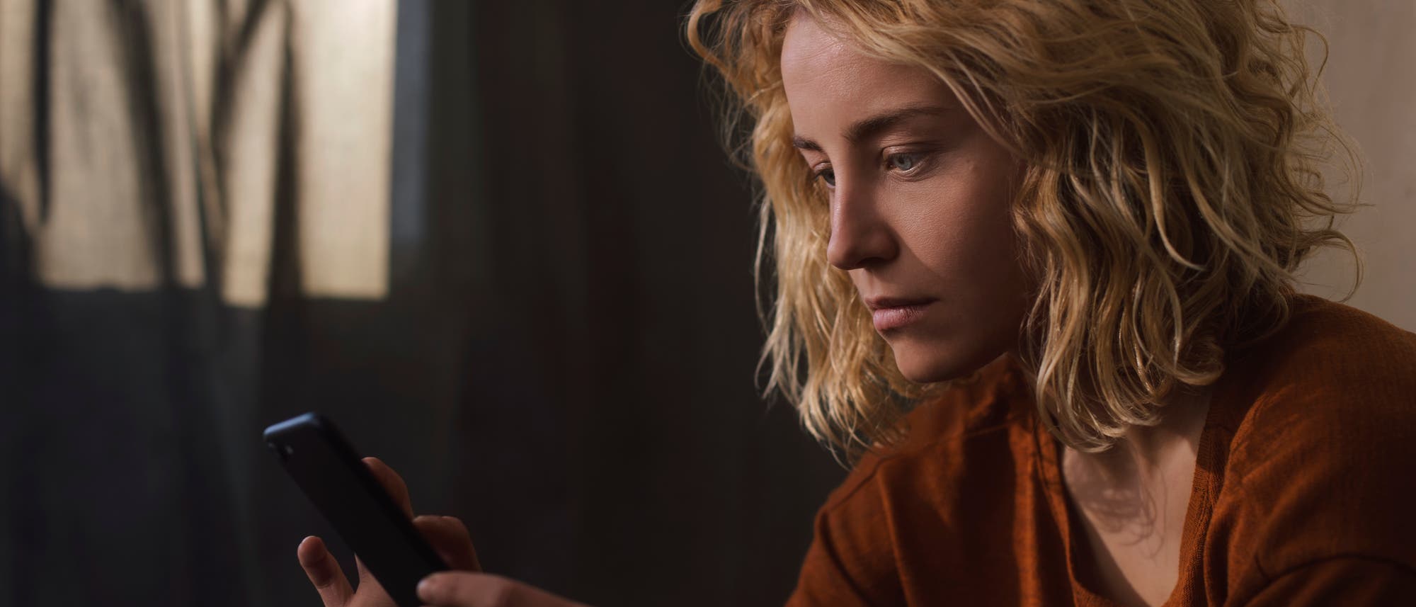 Eine Person sitzt in einem dunklen Raum und schaut auf ein Smartphone. Sie tr&auml;gt ein orangefarbenes Oberteil. Im Hintergrund sind Vorh&auml;nge zu sehen, die weiches Licht durchlassen. Die Szene vermittelt eine nachdenkliche und ein wenig traurige Stimmung.