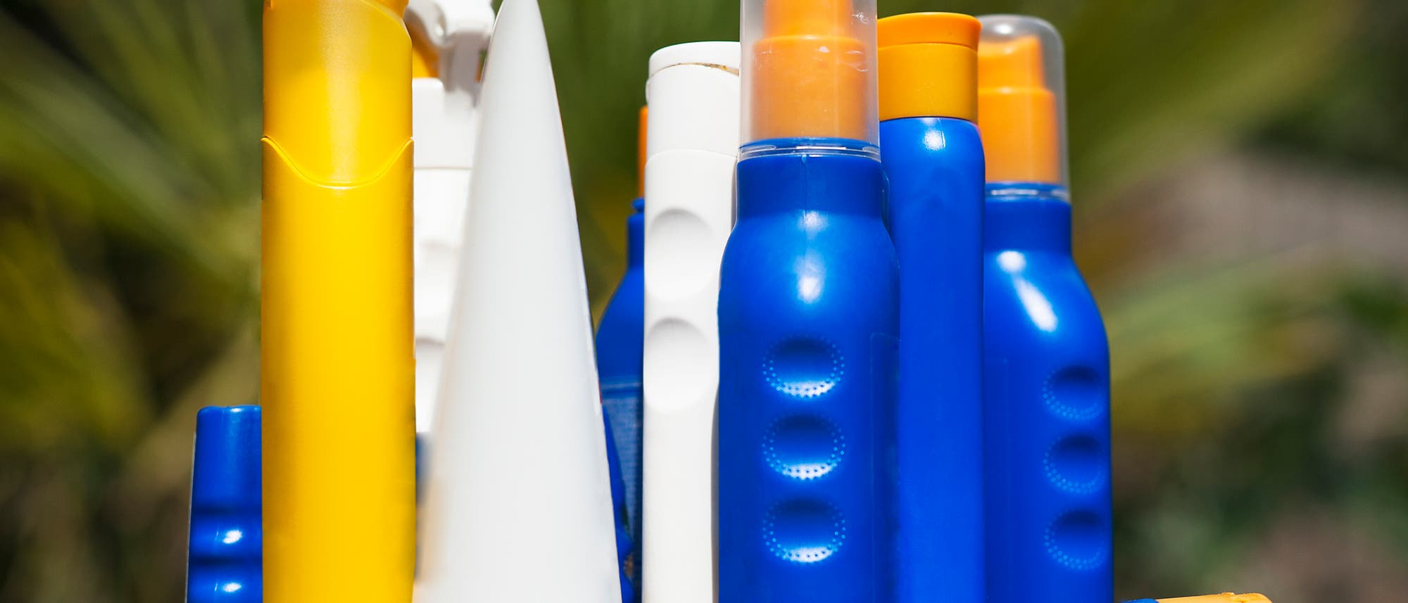 Mehrere bunte Sonnencremeflaschen in Gelb, Blau und Weiß stehen auf einem Tisch im Freien. Die Flaschen sind unterschiedlich geformt und haben orangefarbene Kappen. Im Hintergrund sind unscharfe grüne Blätter zu sehen. Mehrere bunte Sonnencremeflaschen in Gelb, Blau und Weiß stehen auf einem Tisch im Freien. Die Flaschen sind unterschiedlich geformt und haben orangefarbene Kappen. Im Hintergrund sind unscharfe grüne Blätter zu sehen.