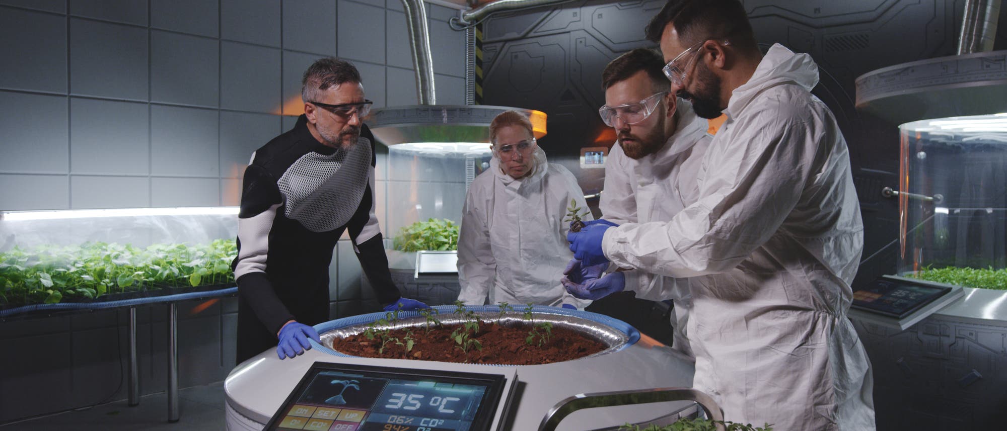 Un grupo de científicos con trajes protectores y gafas examinan plantas en un laboratorio futurista. Están alrededor de una mesa redonda con tierra y plántulas. Una pantalla muestra datos ambientales como la temperatura de 35 grados centígrados. Al fondo se pueden ver otras plantas en contenedores iluminados. La escena transmite una sensación de investigación y tecnología moderna. Un grupo de científicos con trajes protectores y gafas examinan plantas en un laboratorio futurista. Están alrededor de una mesa redonda con tierra y plántulas. Una pantalla muestra datos ambientales como la temperatura de 35 grados centígrados. Al fondo se pueden ver otras plantas en contenedores iluminados. La escena transmite una sensación de investigación y tecnología moderna.