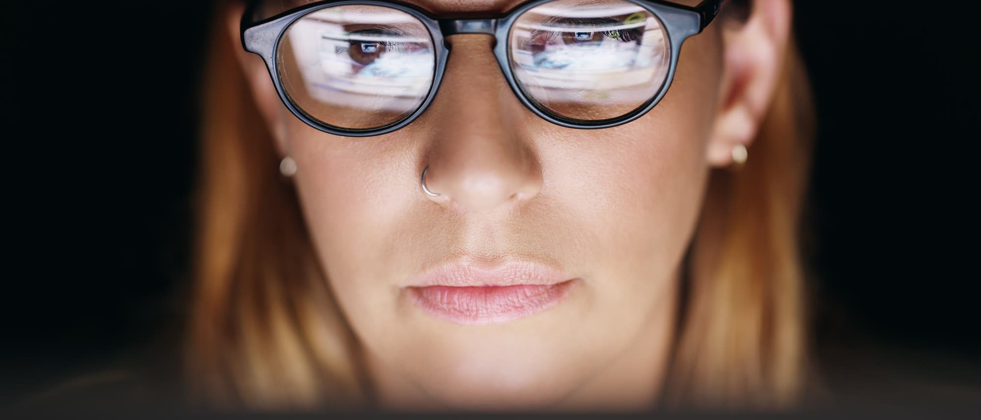 Eine Person mit Brille schaut auf einen Computerbildschirm. Der Bildschirm spiegelt sich in den Brillengläsern wider. Die Szene ist in einem dunklen Raum aufgenommen, was auf konzentriertes Arbeiten oder Surfen im Internet hindeutet.